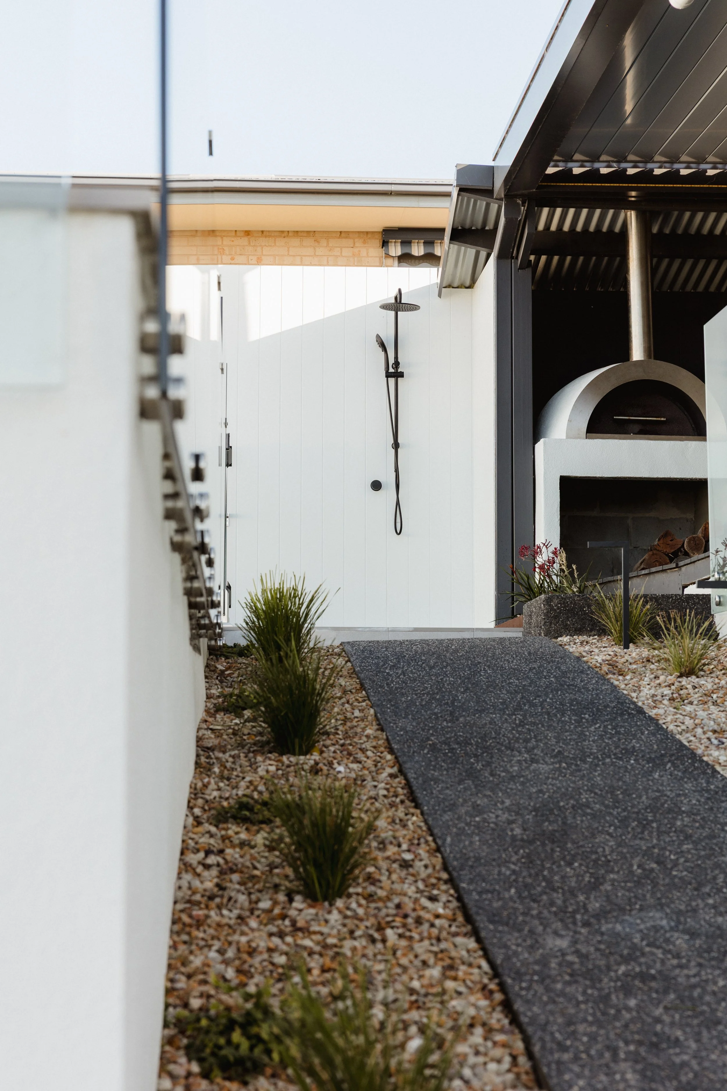 Outdoor shower with rainfall shower head next to an outdoor pizza oven, surrounded by gravel and small plants, with a black ramp leading to the shower