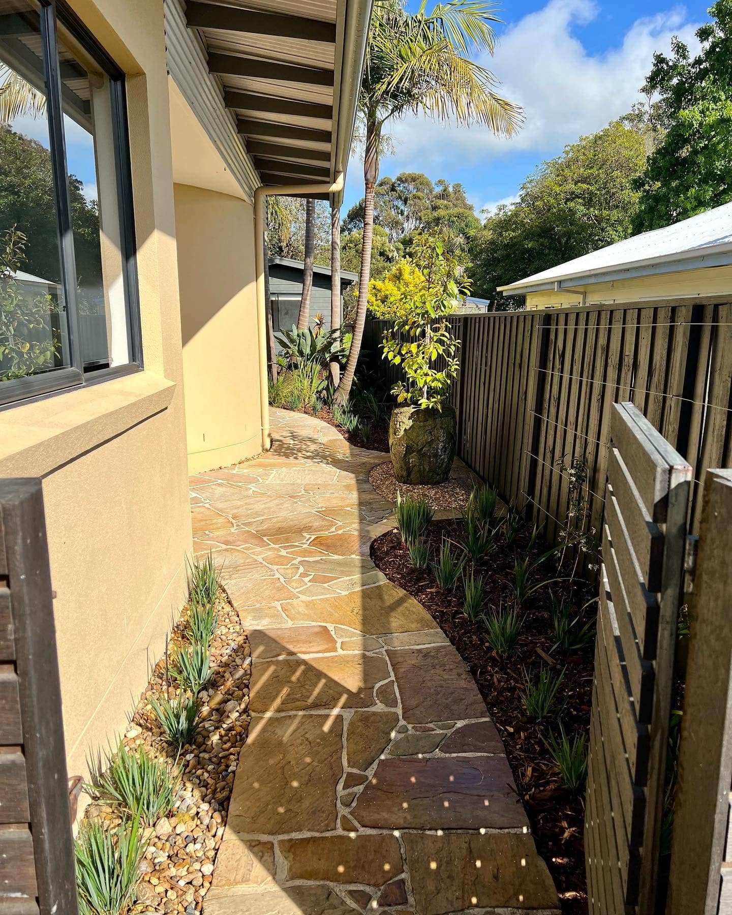 A curved stone pathway in a backyard garden with a beige house on the left, a wooden fence on the right, and tropical plants including palm trees and small bushes.