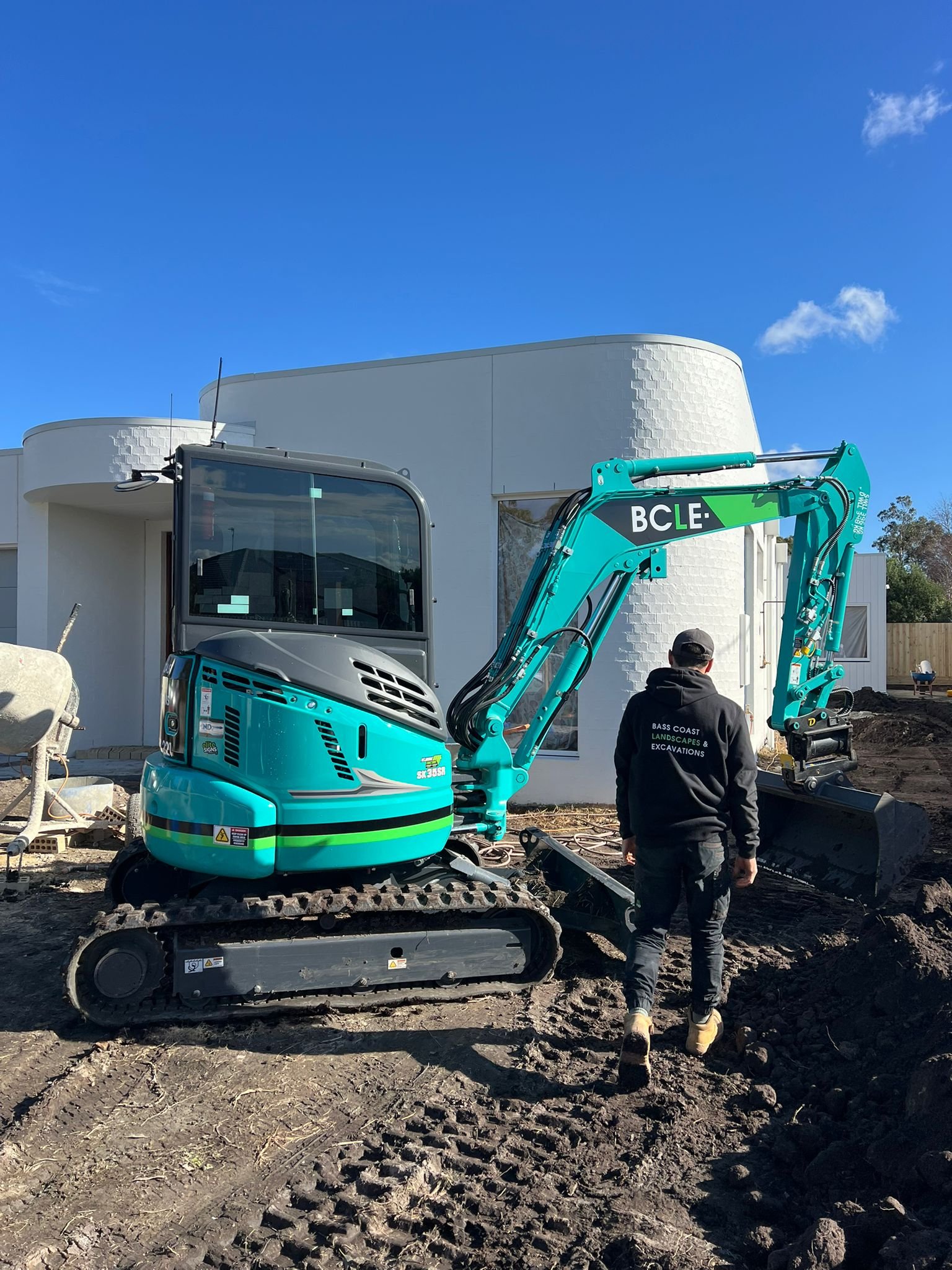 Construction worker walking near a teal mini excavator on a dirt site in front of a white building under a clear blue sky.