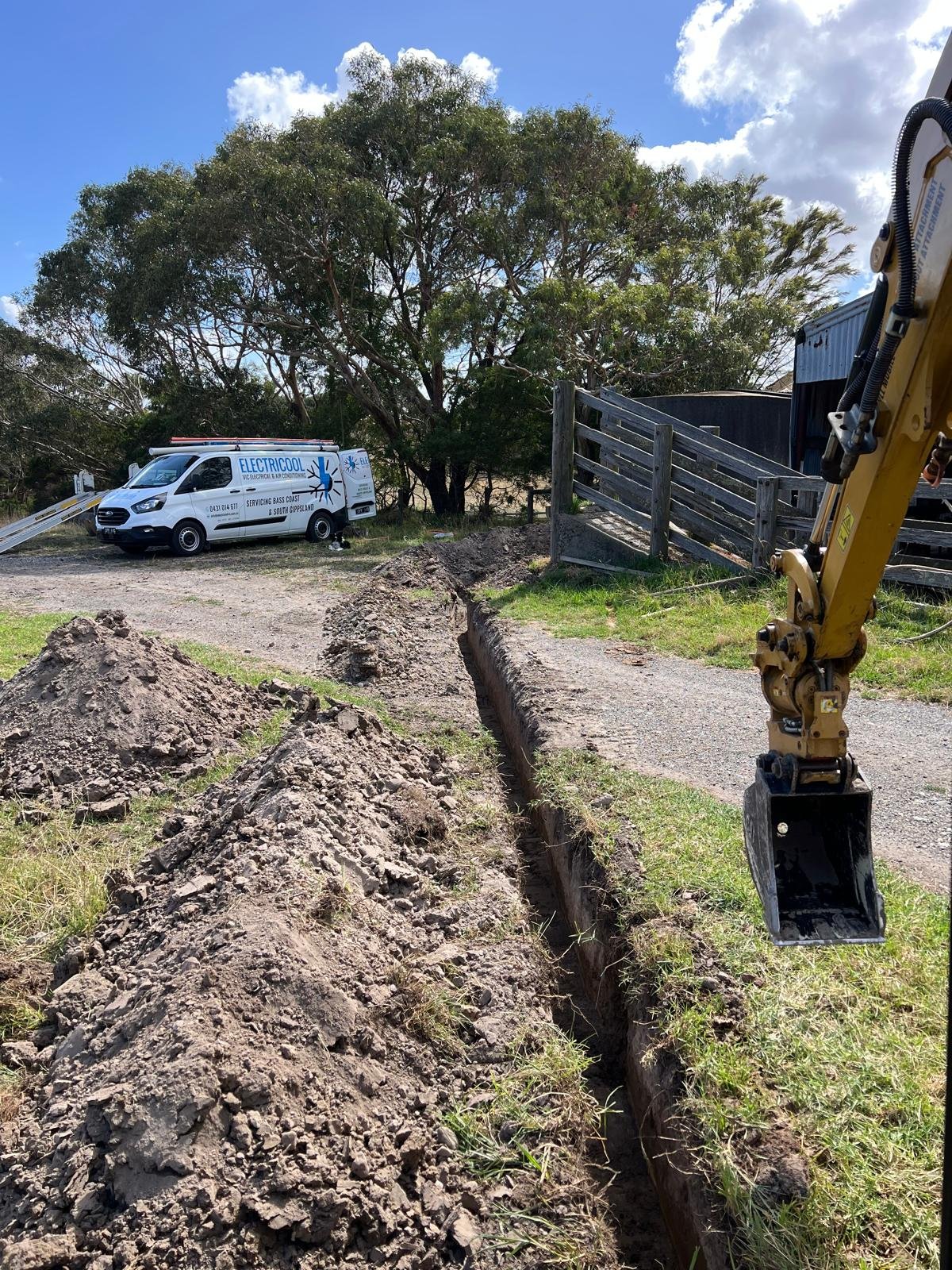 A construction site with a trench in the ground, a yellow excavator's arm, a pile of dirt, a van labeled 'Electric Cool' in the background, and trees under a partly cloudy sky.
