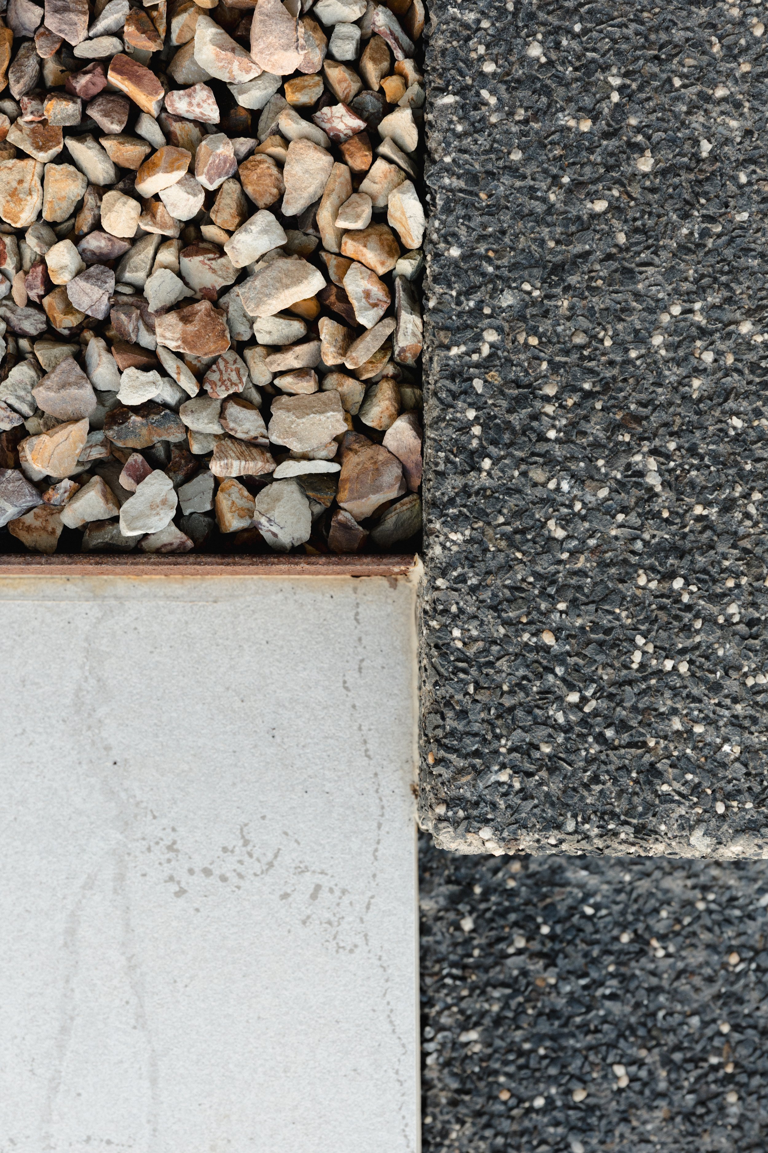 Close-up of four different types of paving materials: small multicolored rocks, black asphalt, large light gray concrete tile, and small black and white gravel.