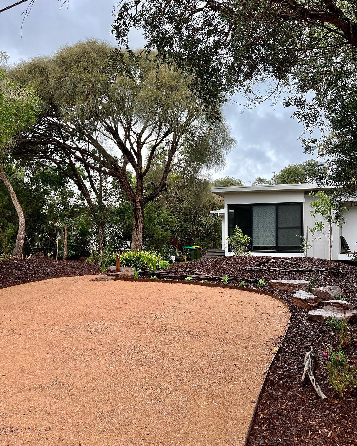 A modern house with large windows and a lush landscape featuring trees, plants, and a well-maintained gravel area.