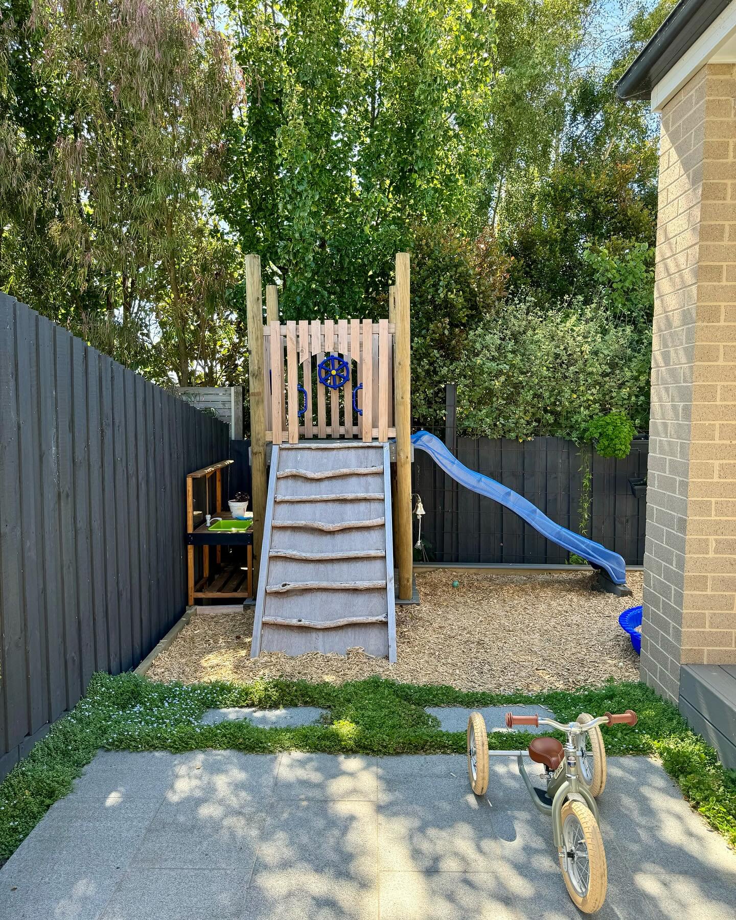 Child's backyard play area with a small slide, climbing structure, and a balance bike on a patio, with trees and a fence in the background.