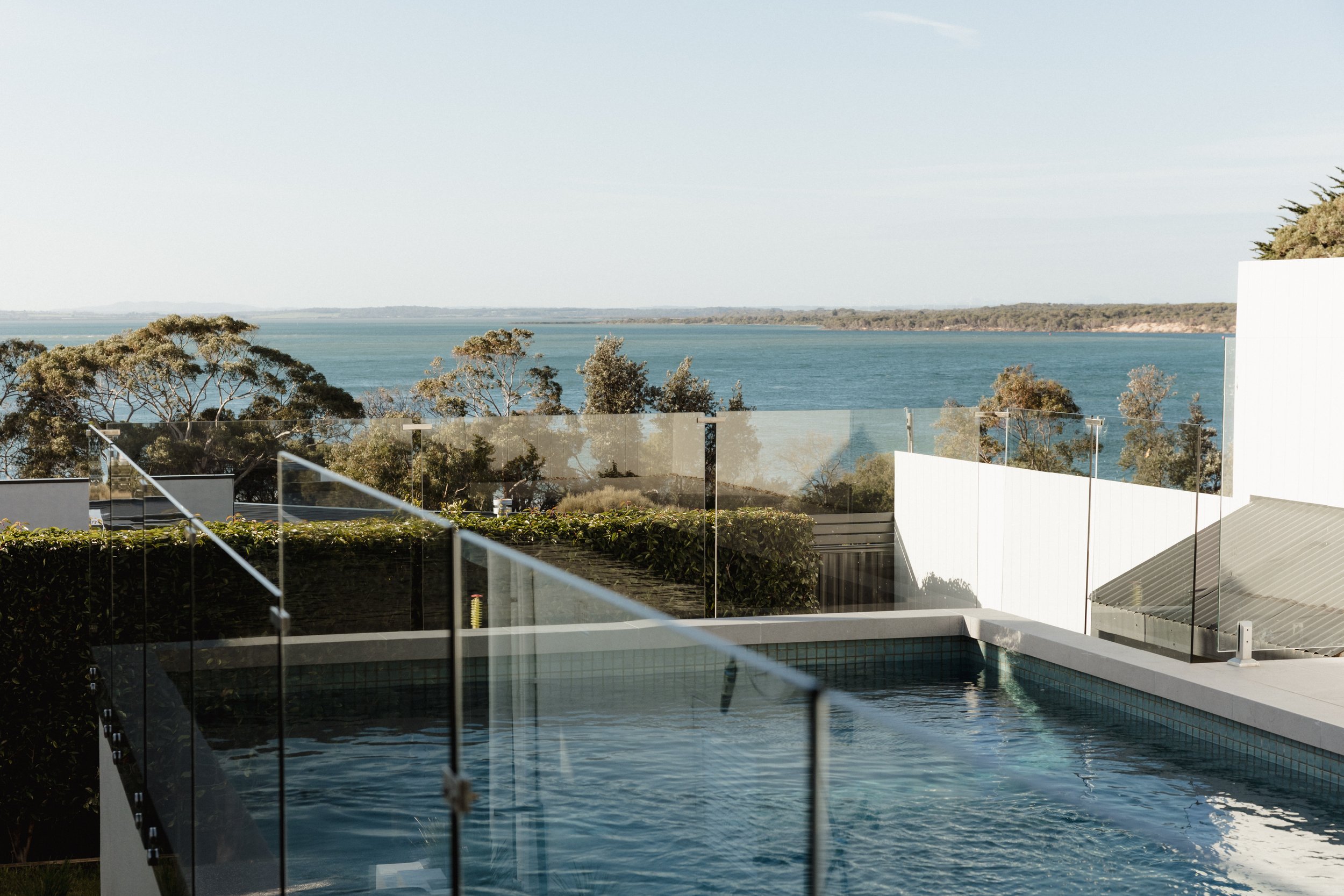 Modern backyard pool with glass safety fencing overlooking trees and a large body of water in the distance under a clear sky.