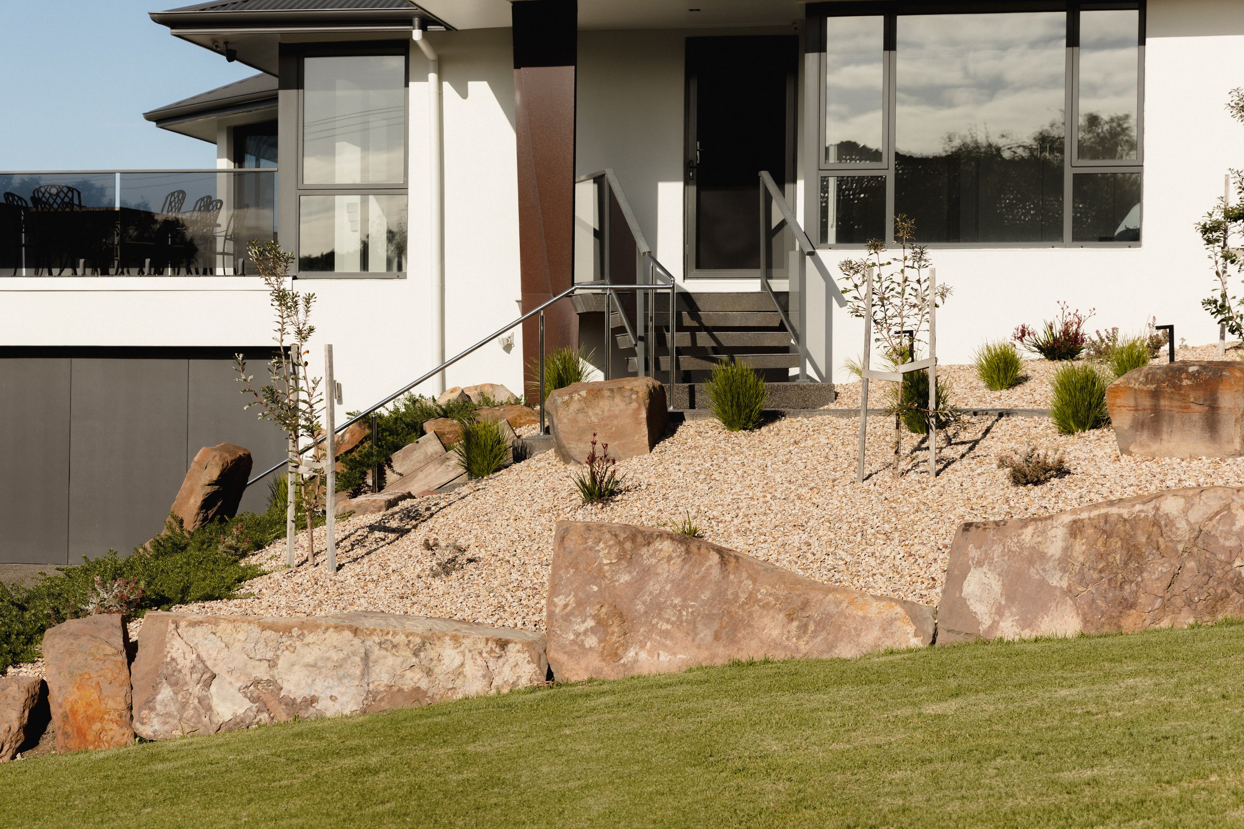 Modern house with large windows, front porch, and landscaped garden with rocks, small trees, and grass.