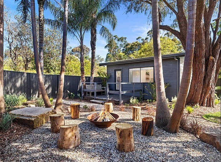 Backyard area with palm trees, a small gray house, and a circular arrangement of tree stumps around a fire pit, gravel ground, and a fenced yard.