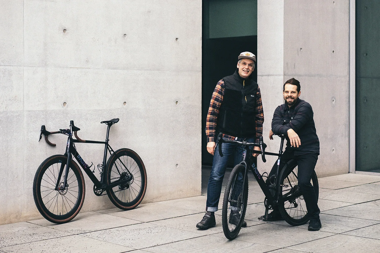 Zwei Männer mit Fahrrädern, eines davon ist ein Elektrofahrrad, stehen vor einer modernen Gebäudewand aus Beton. Das eine Fahrrad ist gegen die Wand gelehnt, das andere wird von einem der Männer gehalten.