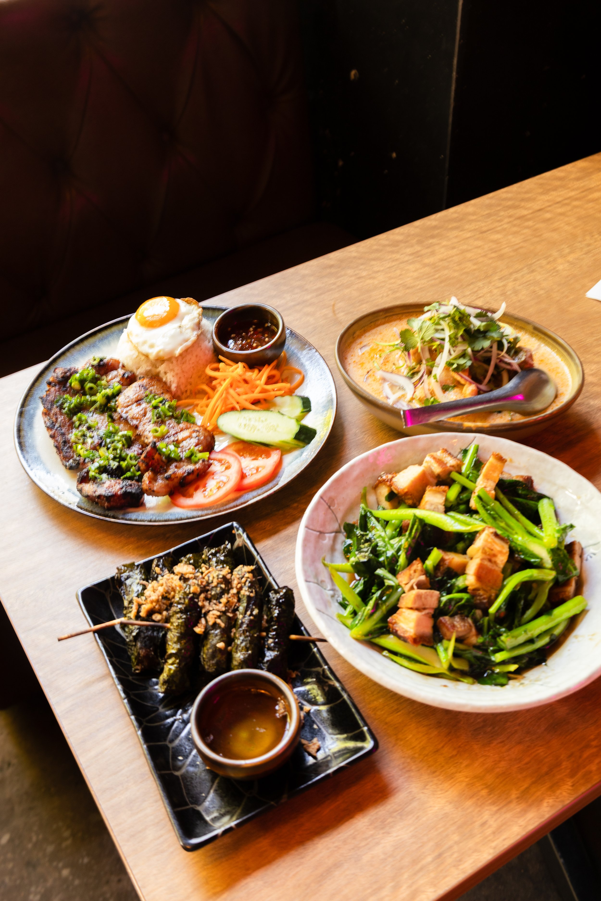A variety of Asian dishes on a wooden table, including a plate with rice, grilled meat, vegetables, and a fried egg, a bowl of curry with vegetables, and a dish with sautéed greens and tofu, served with dipping sauces.