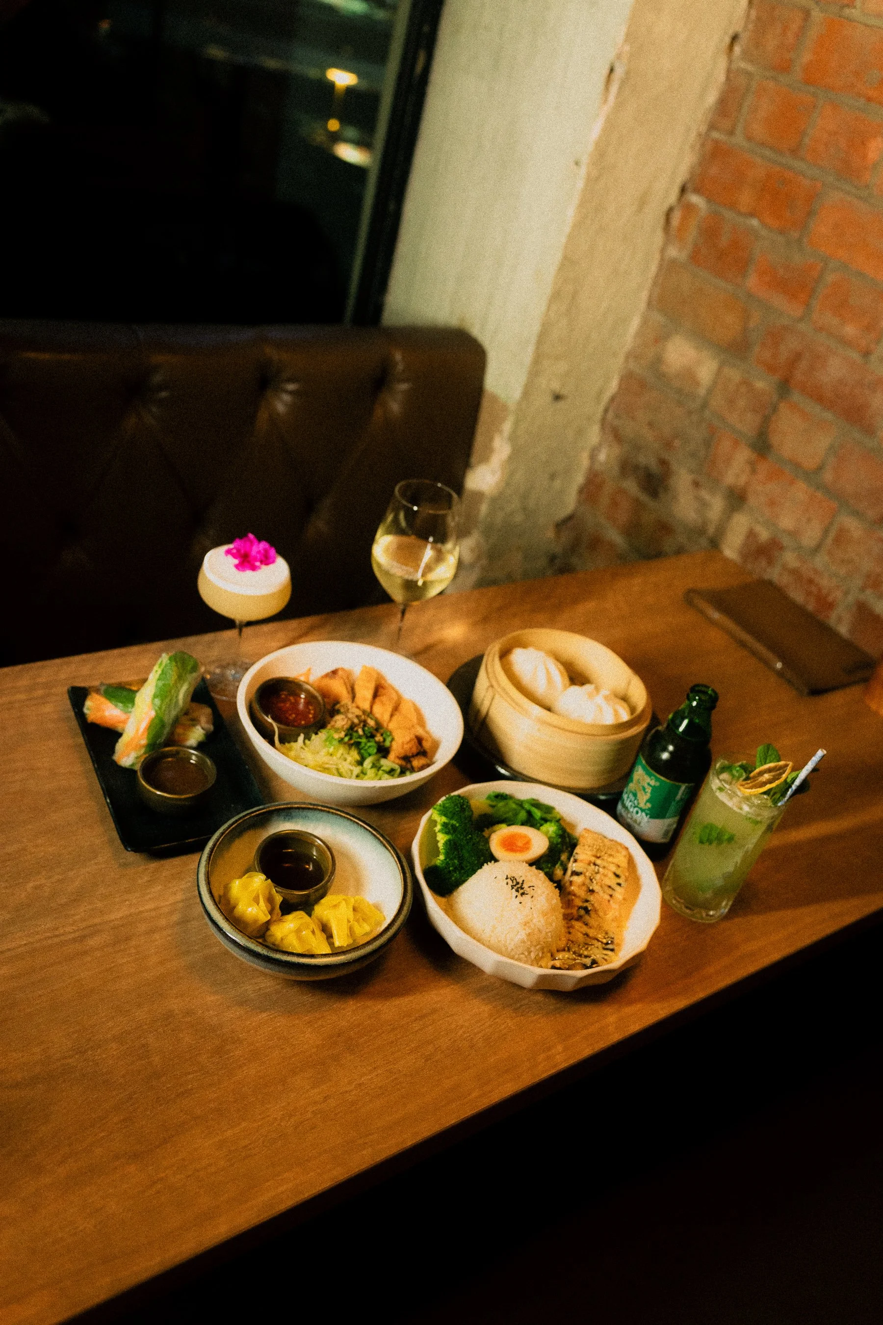 A variety of Asian dishes on a wooden table, including rice, dumplings, sushi rolls, vegetables, and drinks, in a restaurant setting with brick and concrete wall.
