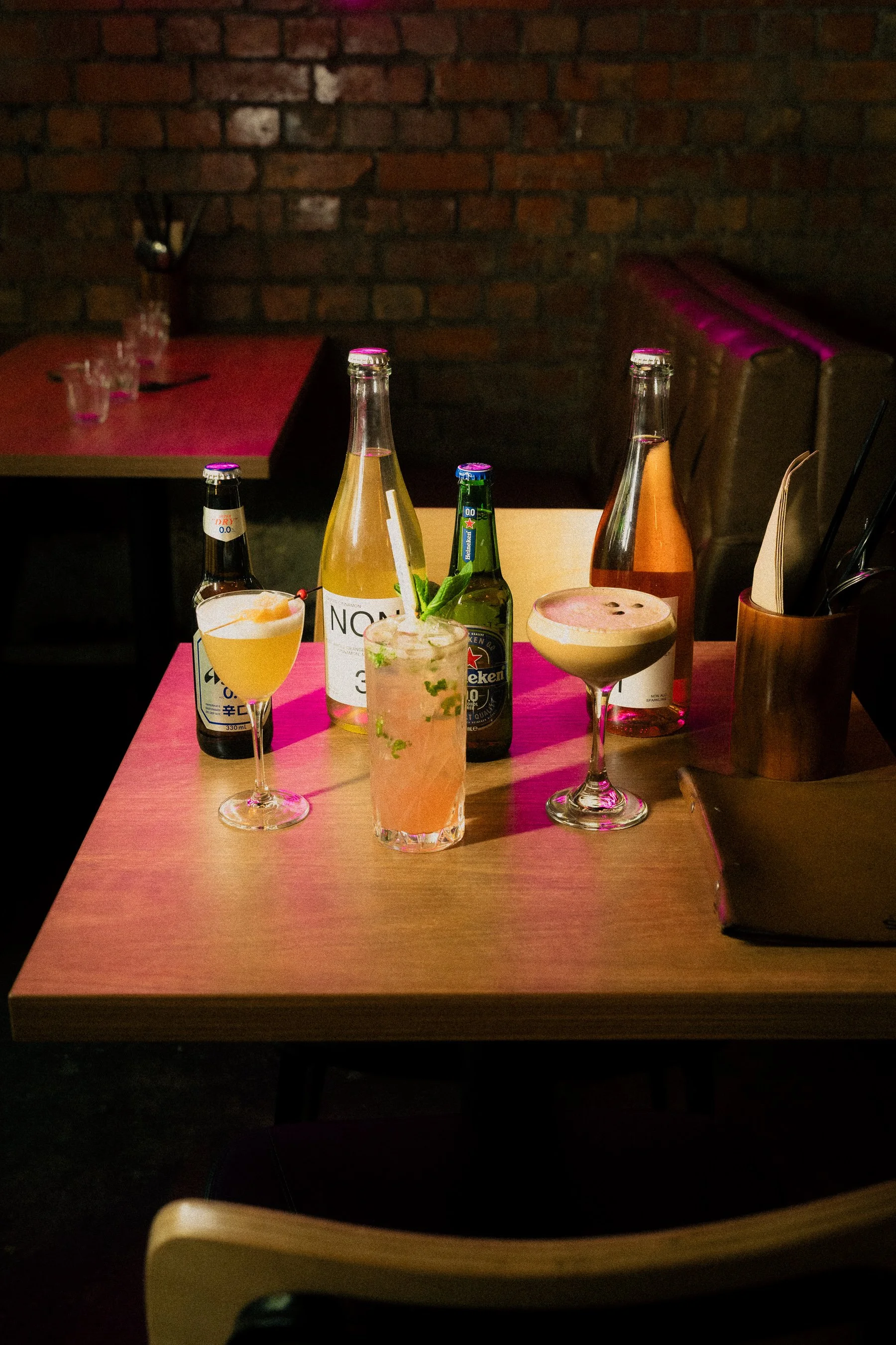 Bar table with six drinks including beers, cocktails, and sodas in various glasses and bottles, next to a wooden container holding utensils, against a brick wall background.