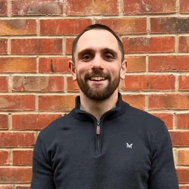 A man with a beard and short hair smiling, standing in front of a brick wall, wearing a black zip-up sweater.