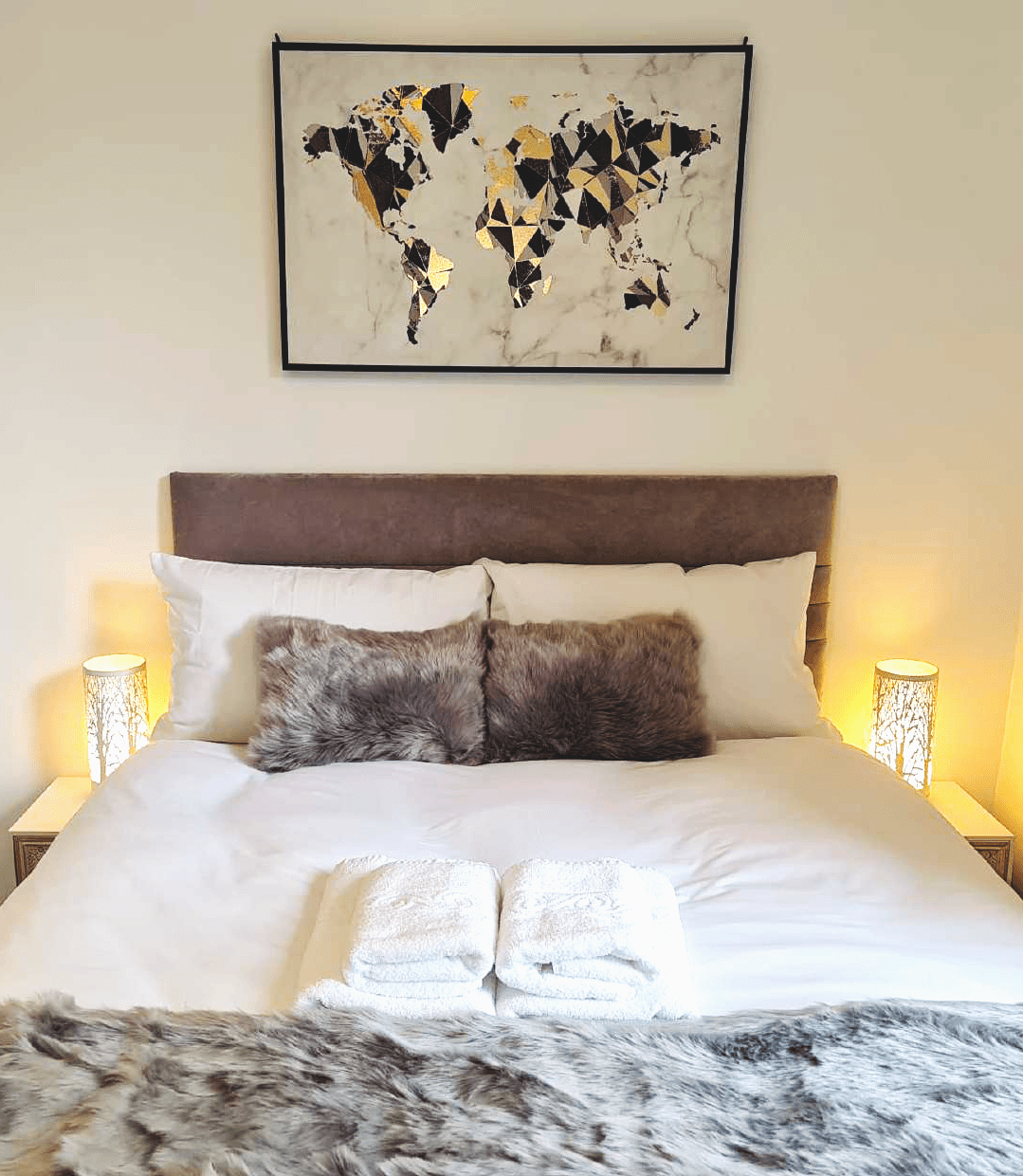 A neatly made bed with two rolled white towels at the foot, fluffy gray pillows, and a fur throw blanket. There are two matching bedside lamps with intricate cut-out patterns on each side of the bed. Above the bed is a framed world map artwork with gold, black, and beige geometric shapes.