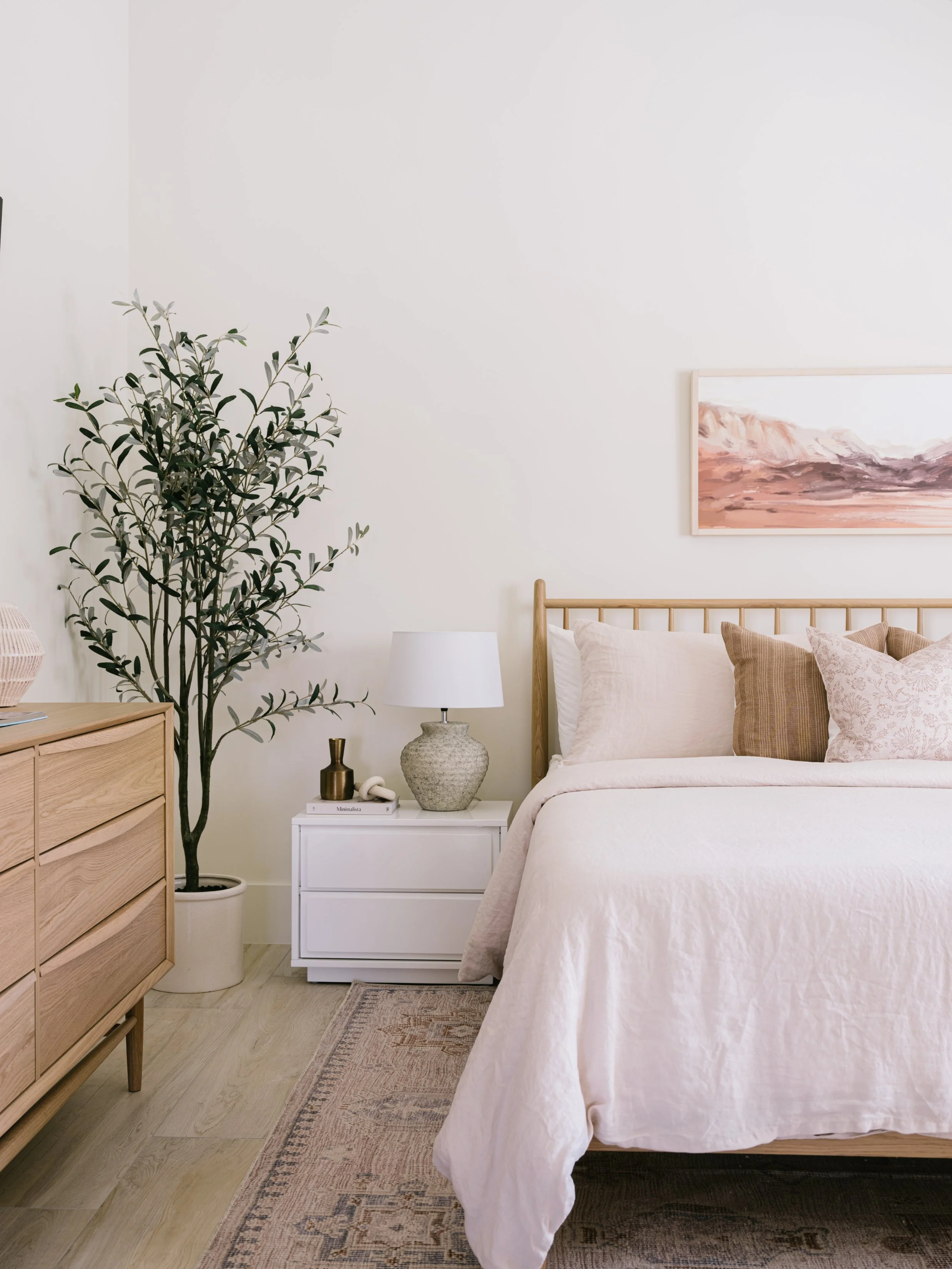 A bedroom with a wooden bed, beige bedding and pillows, a white nightstand with a lamp, a small decorative vase, and a potted plant. A painting hangs on the wall above the bed.