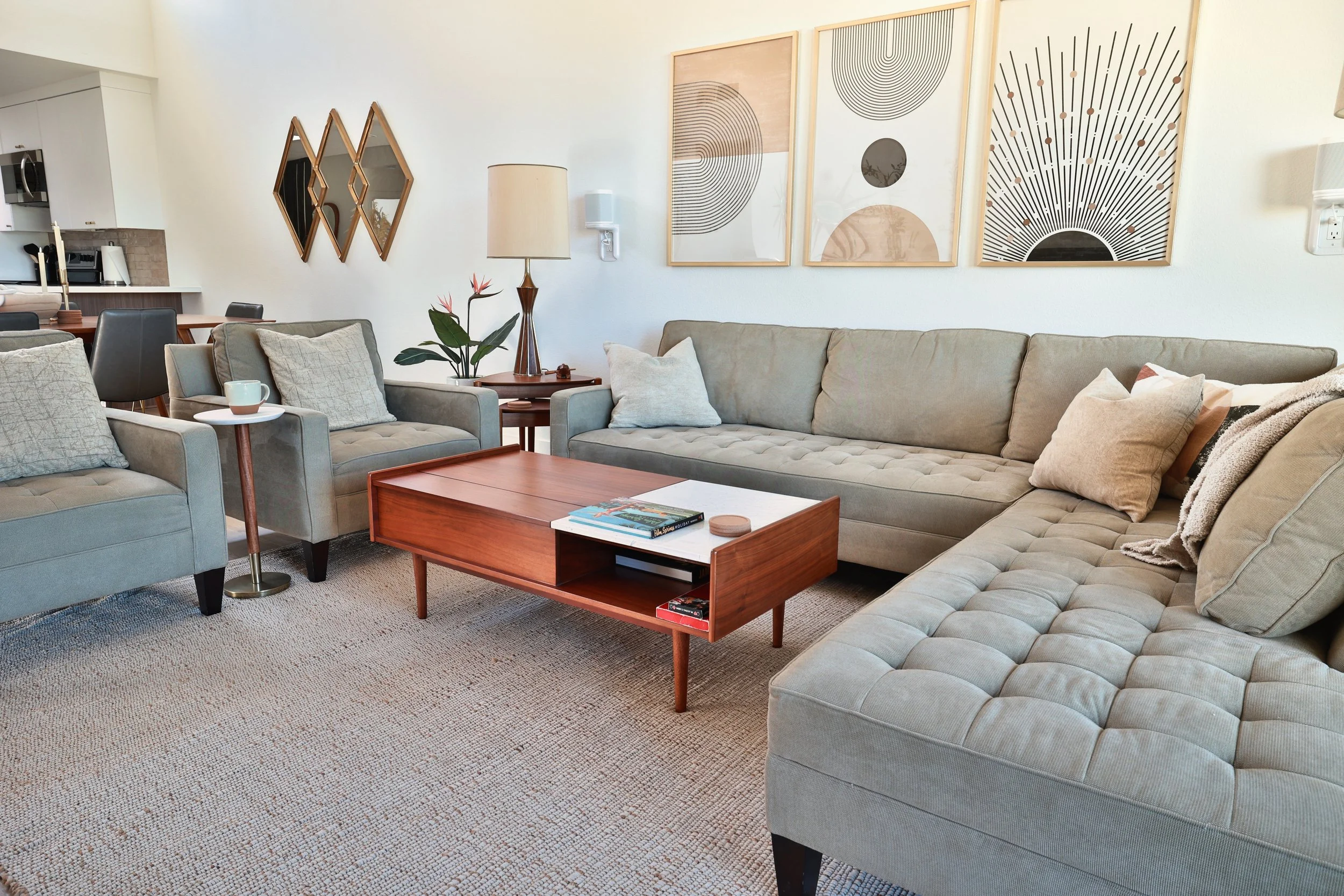 Living room with beige sectional sofa, blue armchair, wooden coffee table, side table with mug, floor lamp, wall art, and a plant in a pot. Background shows kitchen and dining area.