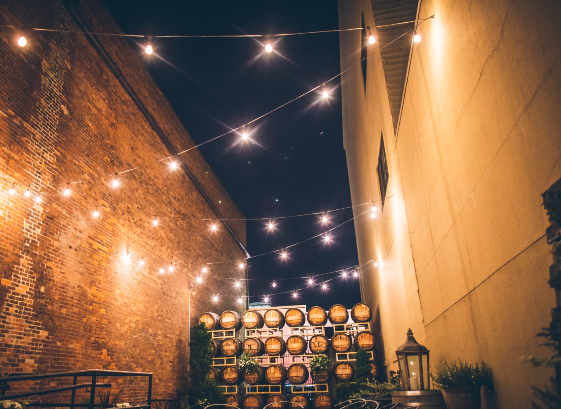 Night scene in an outdoor alley with string lights hanging overhead, brick wall on the left, beige wall on the right, and stacks of wine barrels at the end with potted plants and lanterns.