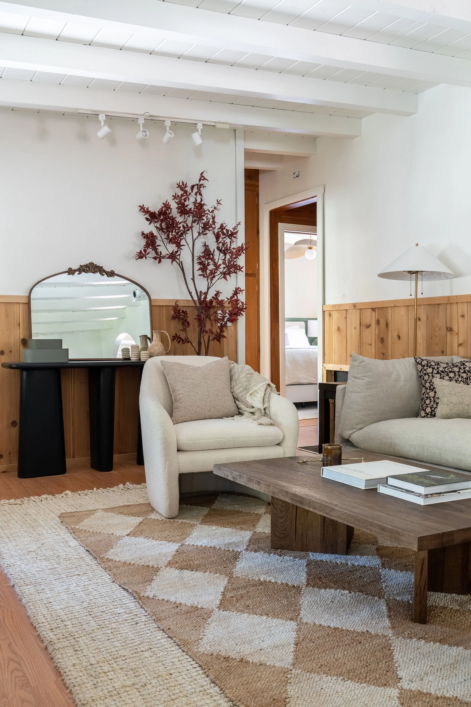 Living room with beige sofa, patterned throw pillows, wooden coffee table with books and candles, armchair, wall-mounted lamp, mirror, decorative red plant, and a doorway leading to a bedroom with a bed and ceiling fan.