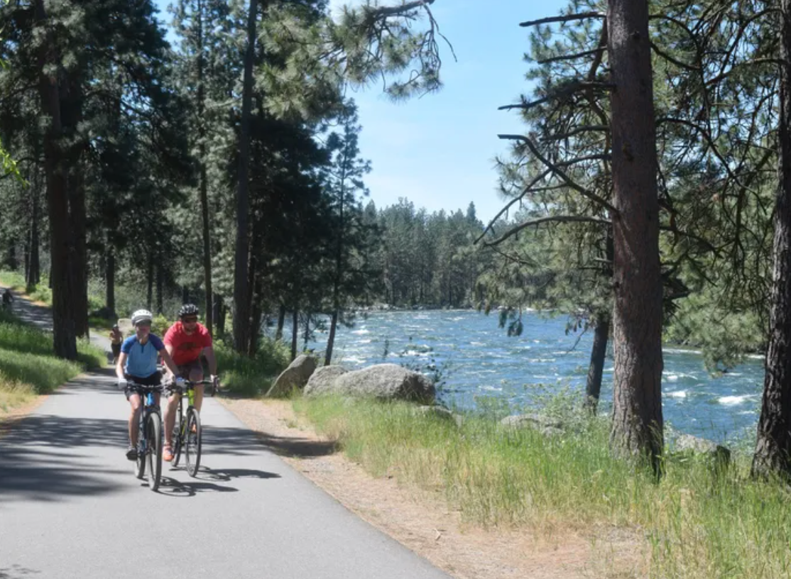 Two children riding bicycles along a forested path next to a river