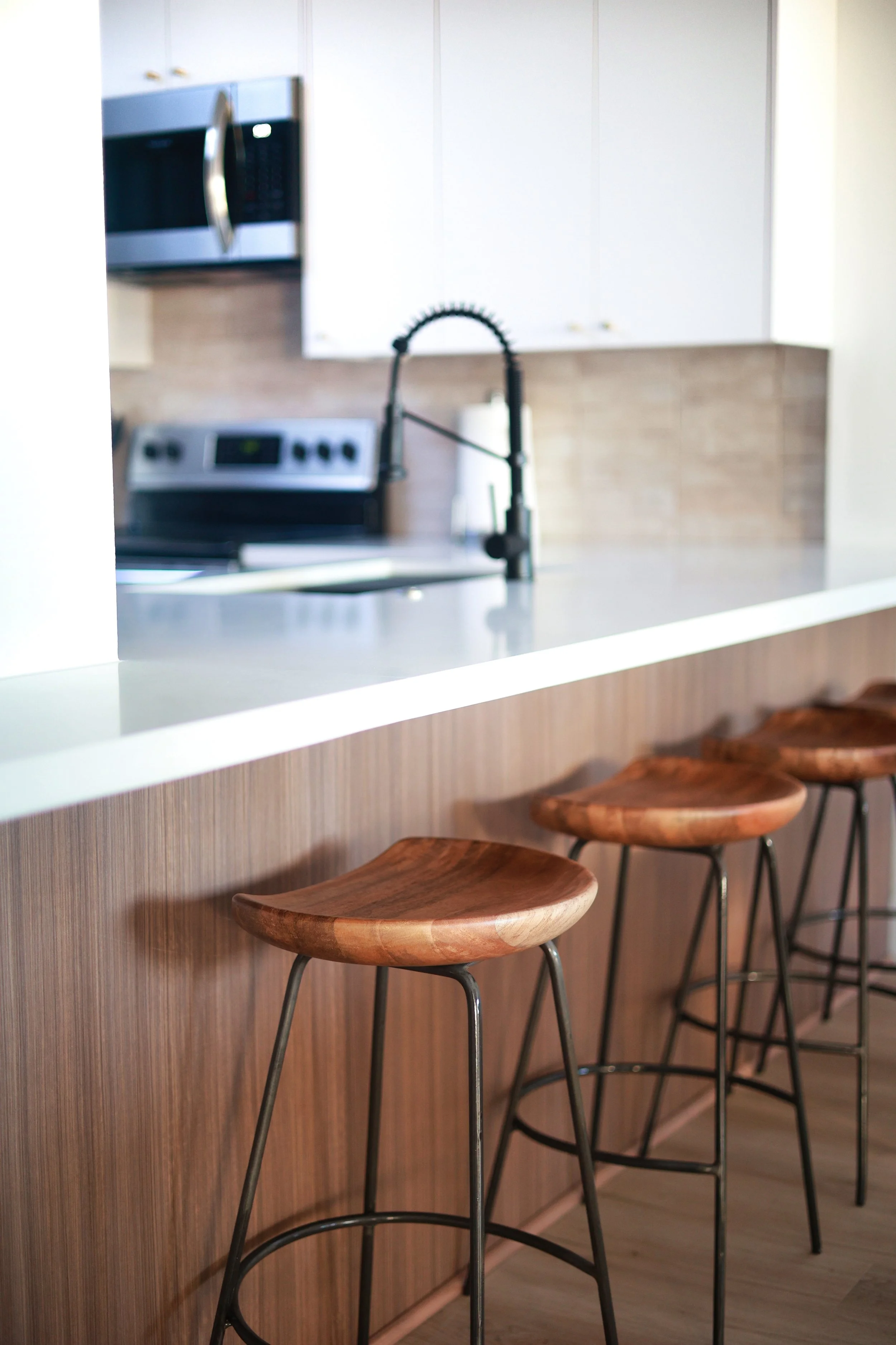 A modern kitchen with white cabinets, a black faucet, a range with an oven, and a microwave. Wooden bar stools are positioned at a white countertop with a wood-paneled base.