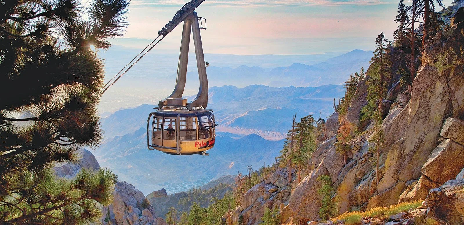 A cable car descending from a mountain with rocky terrain and forested slopes, overlooking a valley with mountain ranges in the distance at sunset.