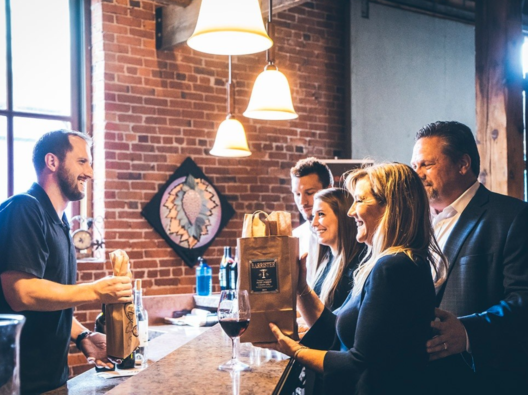 Group of people at a bar receiving gifts from the bartender, with a brick wall and artwork in the background.