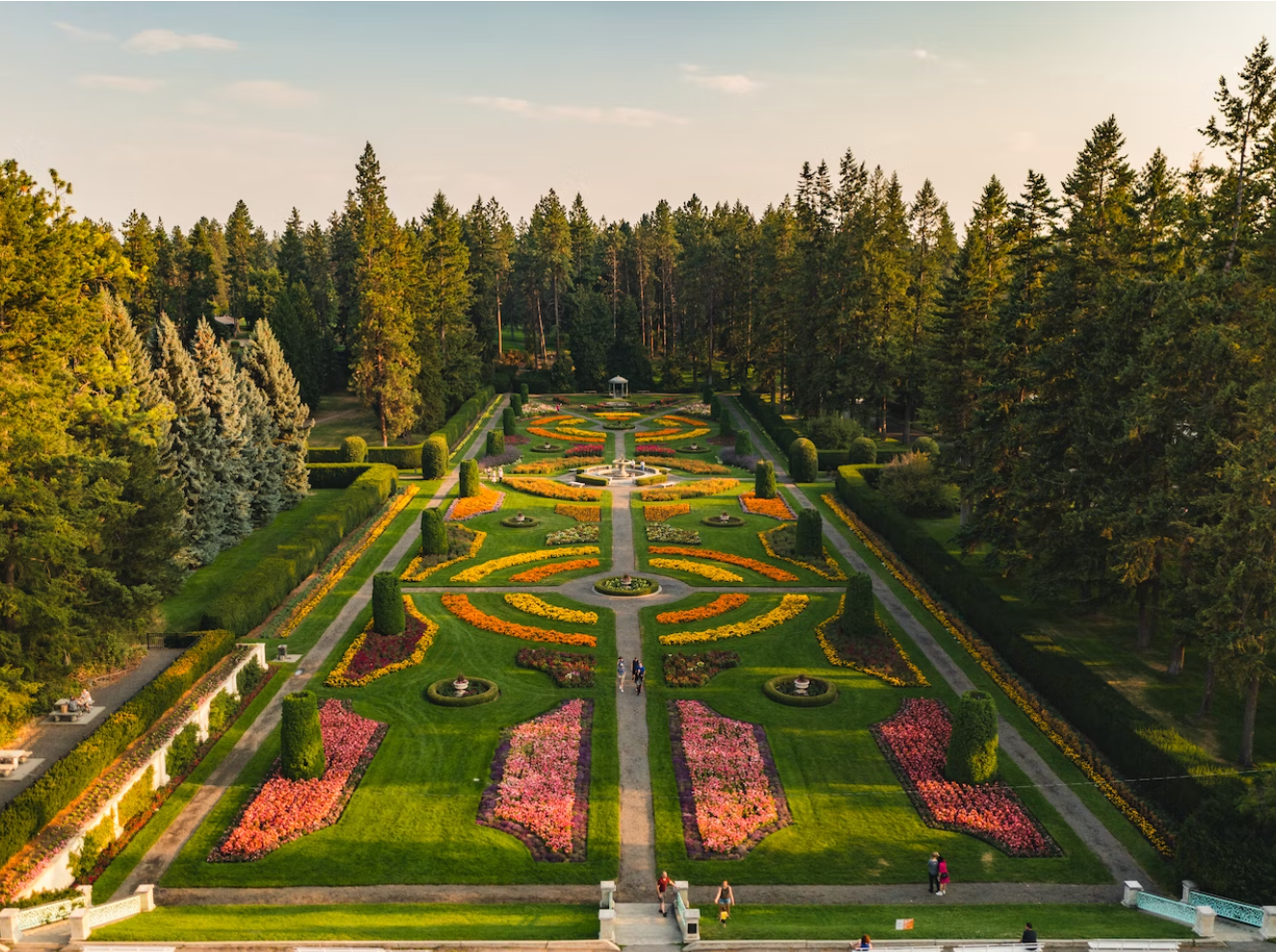 A beautifully landscaped formal garden with colorful flower beds, green lawns, and tall trees surrounding it.