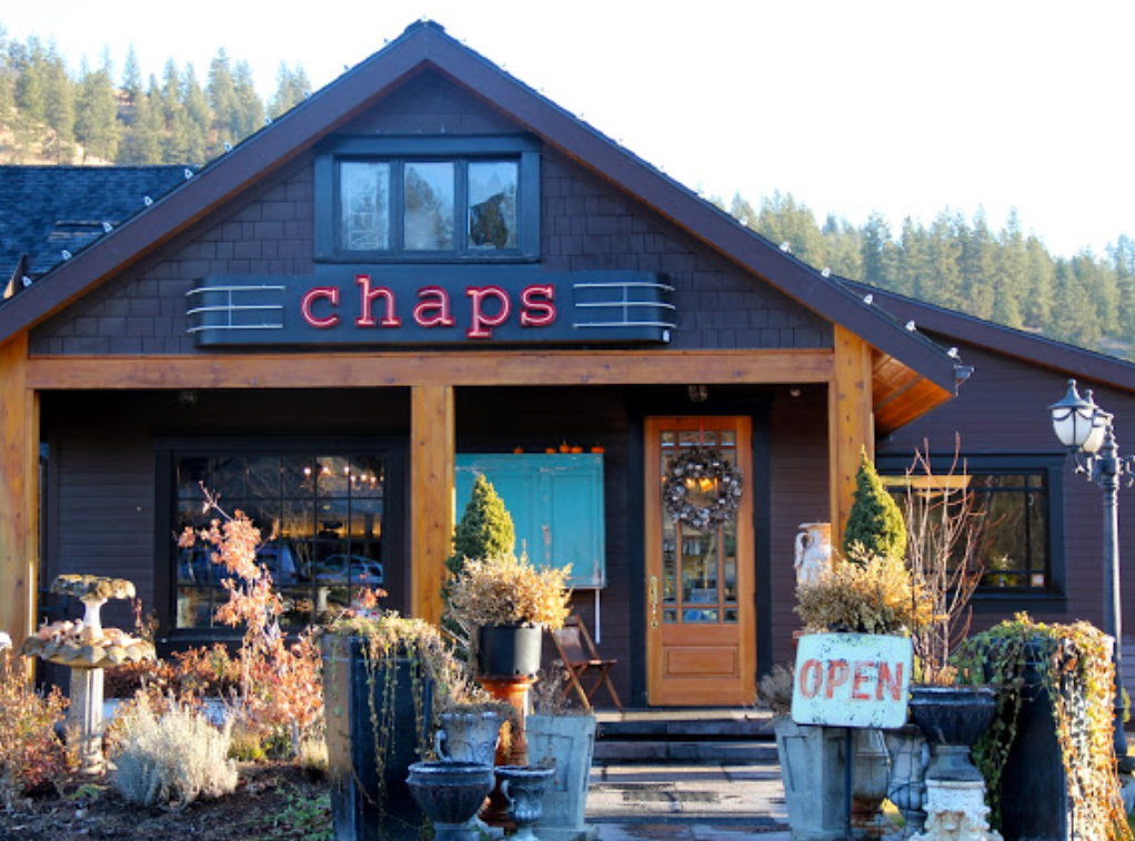 Front view of a rustic shop named 'chaps' with a wooden door, decorated for autumn, outside display with plants, pumpkins, an 'Open' sign, and surrounded by outdoor lanterns, set against a forested background