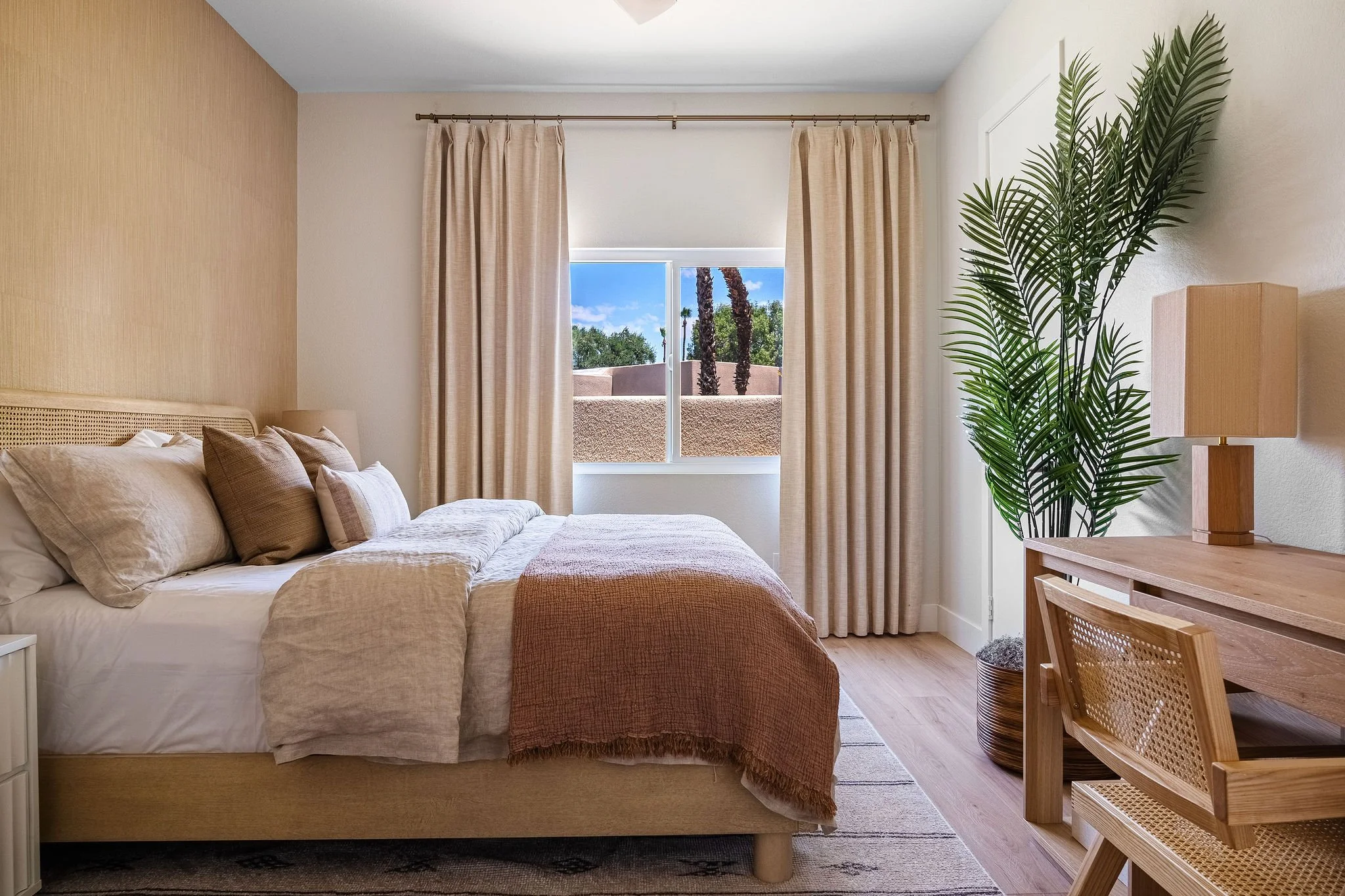 A bedroom with a bed, beige bedding, multiple pillows, a large window with beige curtains, a potted plant, a wooden desk with a lamp, and wooden furniture.