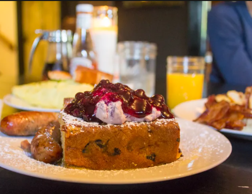 A plate of French toast topped with whipped cream and cherry sauce, served with sausage and sides, breakfast scene.