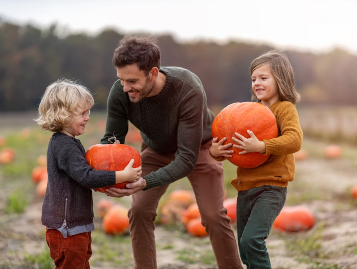 A father and two children in a pumpkin patch, holding large pumpkins and smiling at each other.