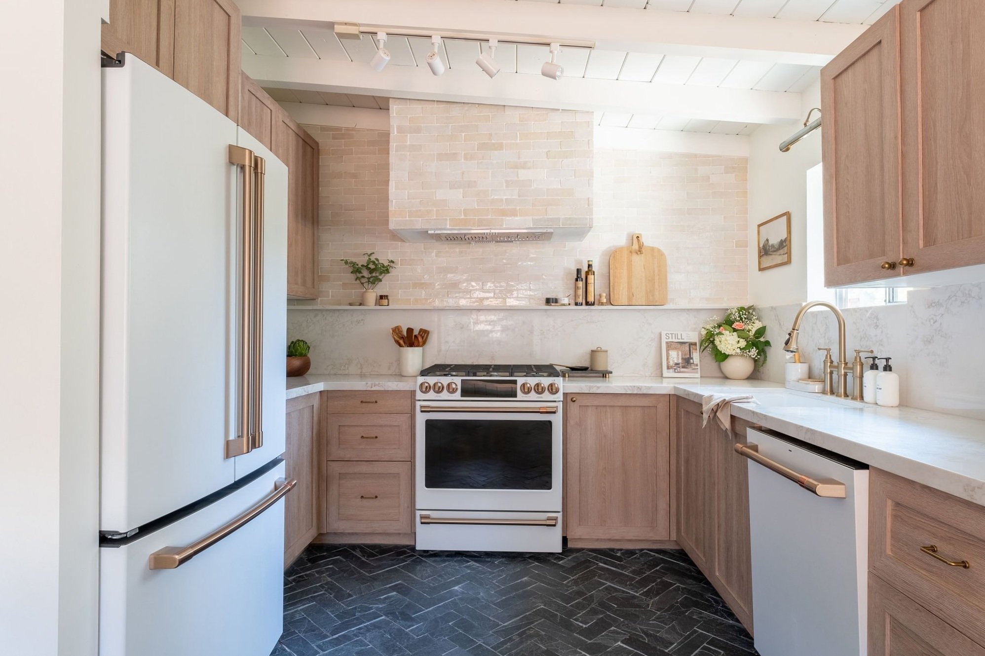 Kitchen with light wood cabinetry, white marble countertops, a white stove with brass accents, a white refrigerator, black herringbone patterned floor, and a white brick backsplash with a range hood. Decor includes potted plants, cutting boards, framed artwork, and soap dispensers.
