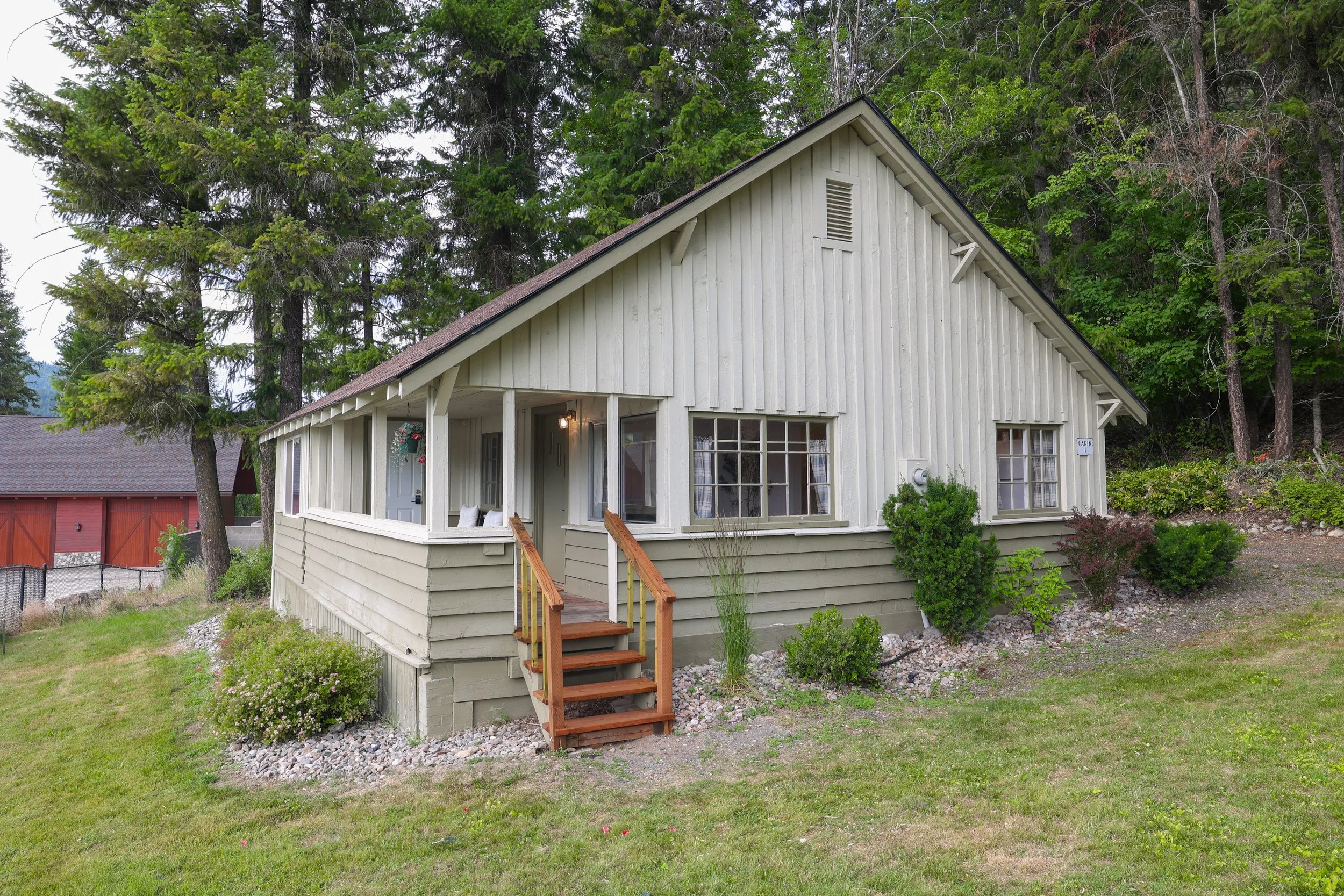 A white wooden house with a front porch has a backdrop of tall green trees and a sloped lawn with bushes and flowers.