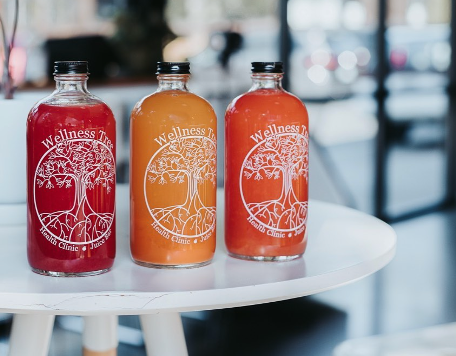 Three bottles of colorful juice on a white table inside a health clinic or juice bar, labeled 'Wellness Tree' with a tree logo.
