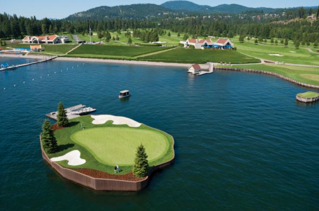 A scenic aerial view of a golf course with a water hazard and a small island green, surrounded by lush green grass, trees, and a building in the background.