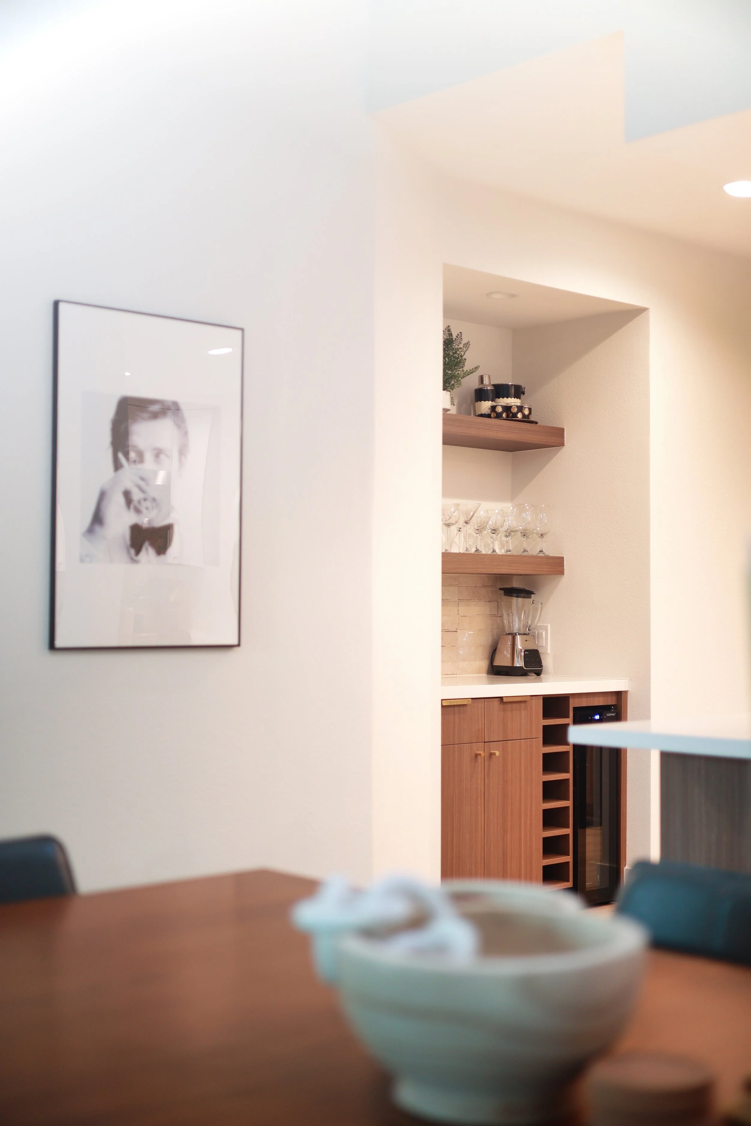 A part of a dining area with out-of-focus bowls on a wooden table in the foreground and a white wall with modern art behind it. There's a small hidden bar area with wooden shelves, glassware, a blender, and a wine fridge.