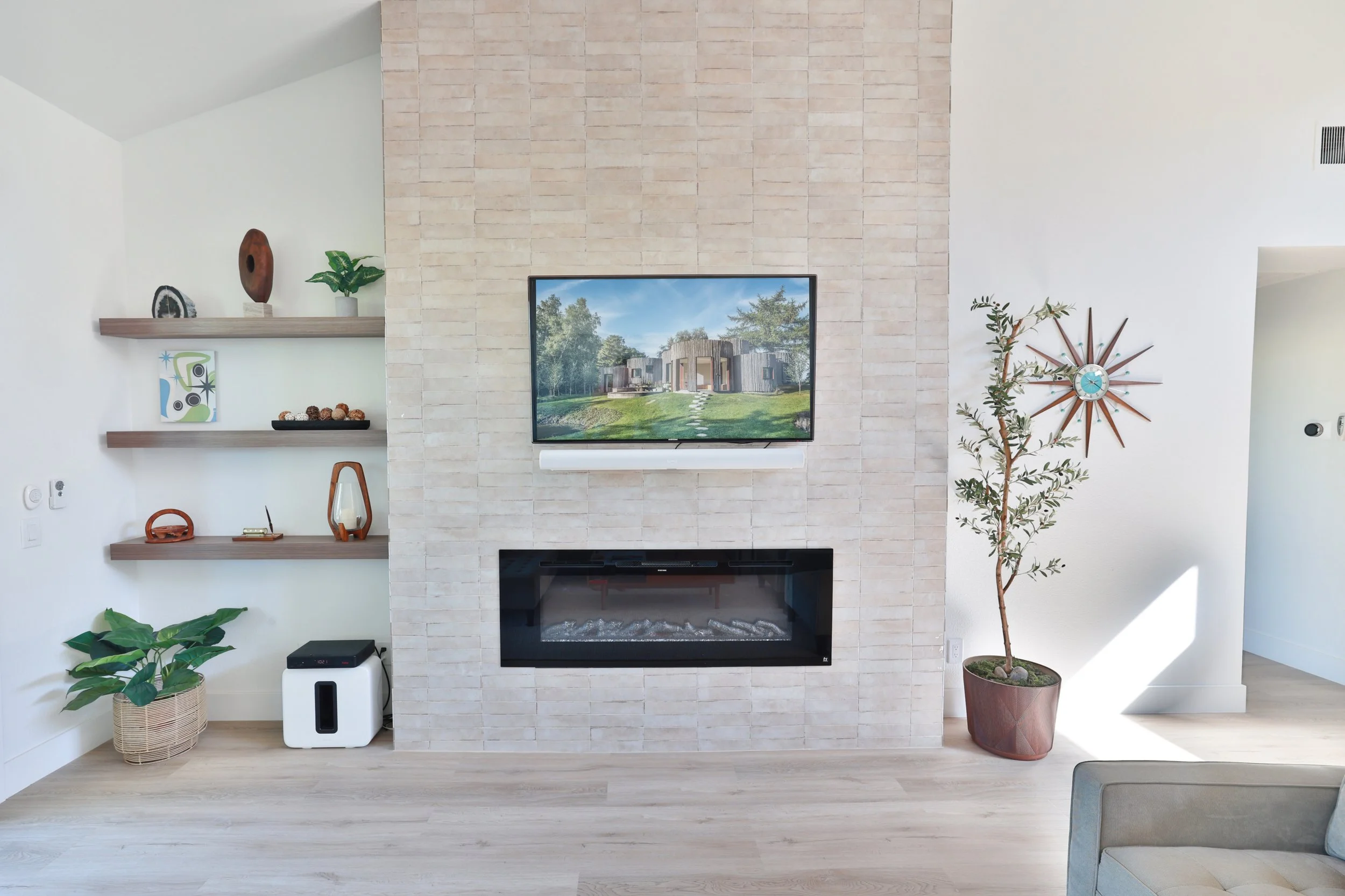 Living room with a modern fireplace, mounted TV above, beige brick wall, and wooden shelves with decorative items. Potted plants on the floor and a wall clock with rays on the right.
