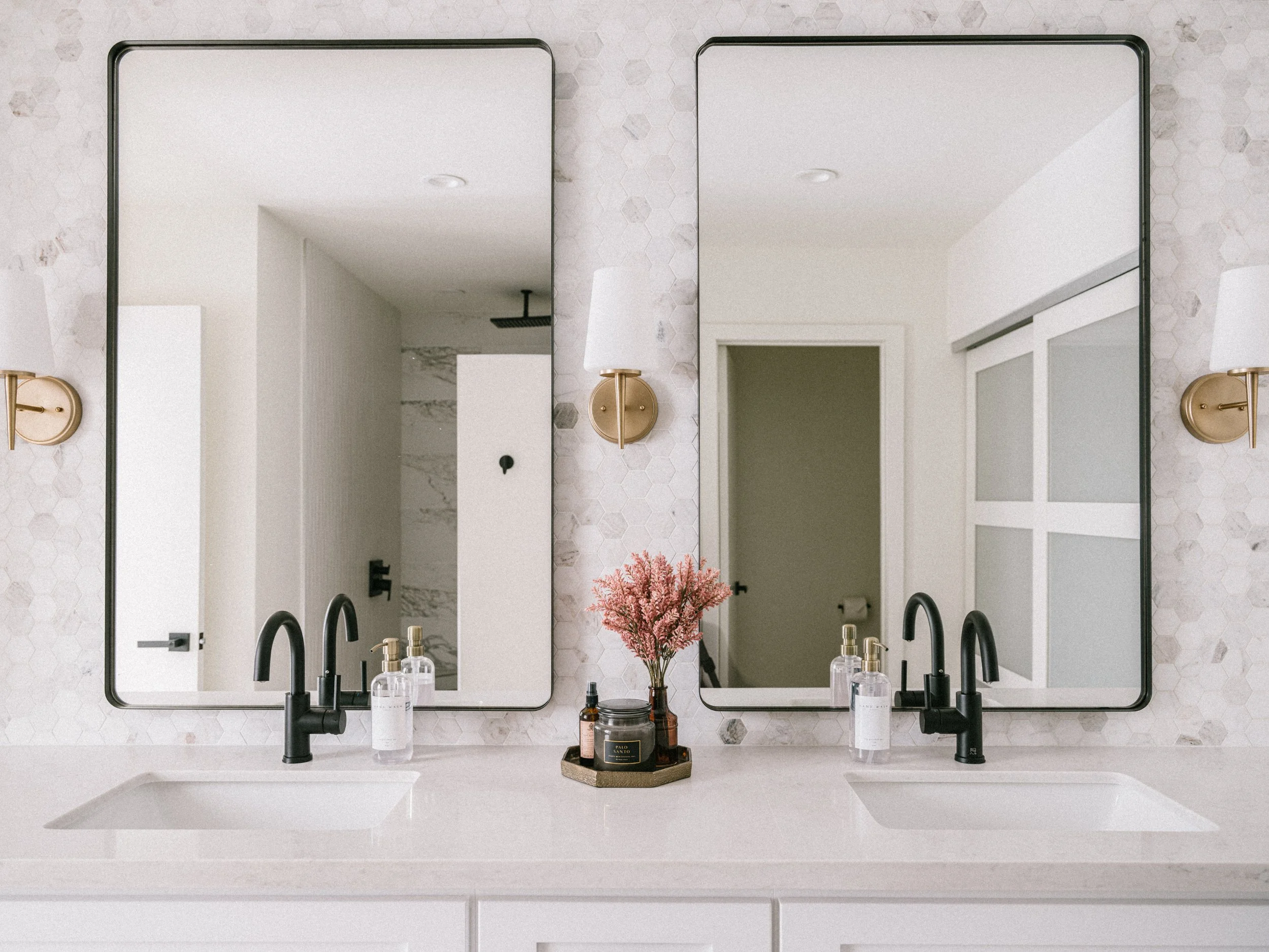 Bathroom with two large mirrors, black faucets, toothbrush holders, a pink flower arrangement, and toiletries on a white countertop.