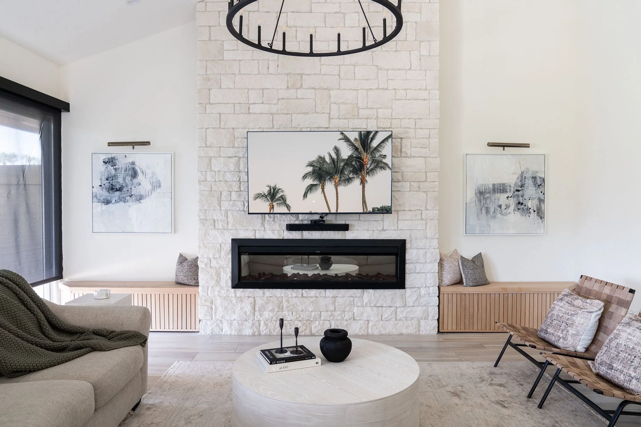 Modern living room with white brick fireplace, wall-mounted TV displaying palm trees, abstract artwork, beige sofa with blanket, wooden benches with pillows, and round coffee table.
