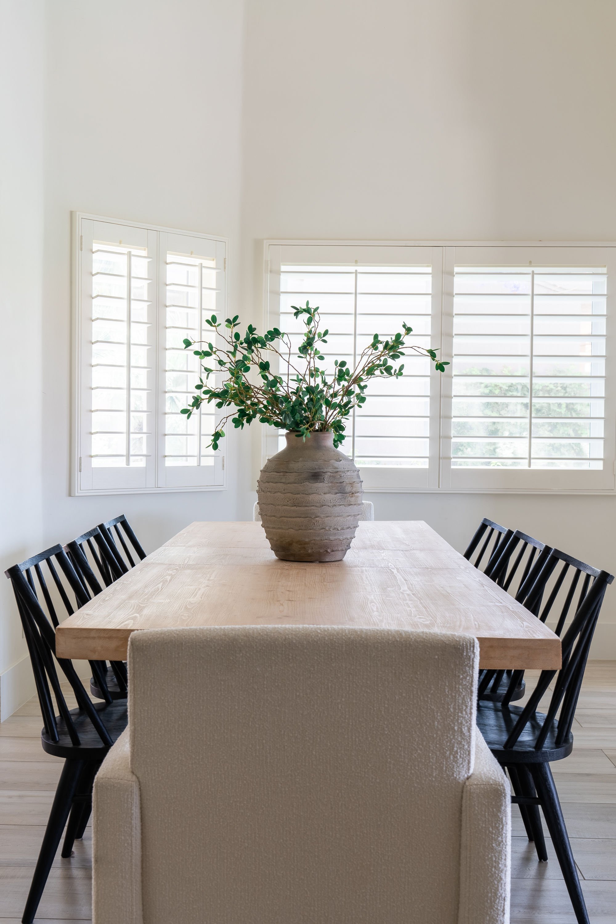 Modern dining room with a wooden table, black chairs, a beige upholstered armchair at the end, large window shutters, and a large potted plant on the table.