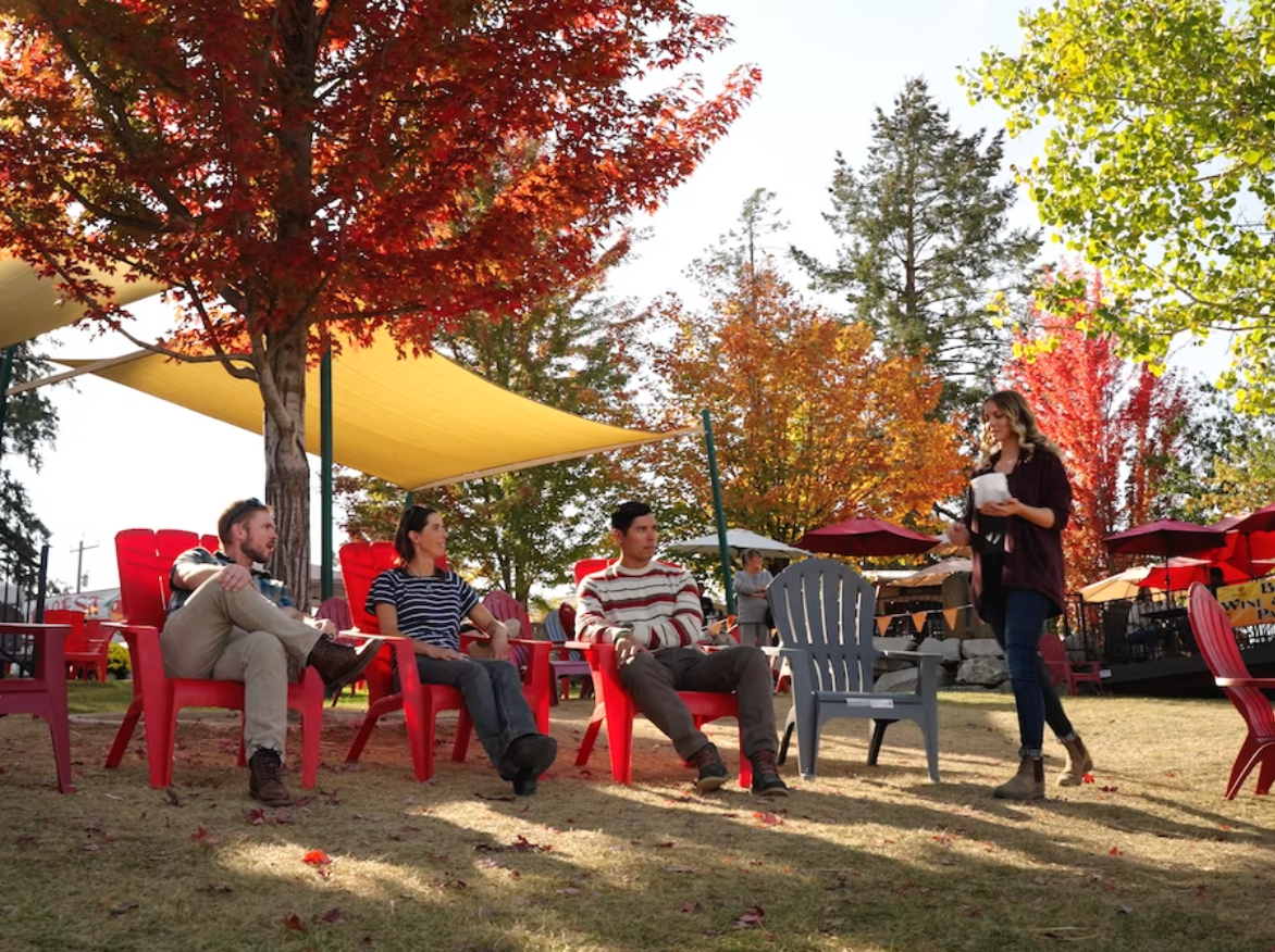 Four people sitting in red and gray outdoor chairs under a tree with colorful autumn leaves, listening to a woman standing and speaking, in an outdoor setting with sun shade sails and umbrellas.