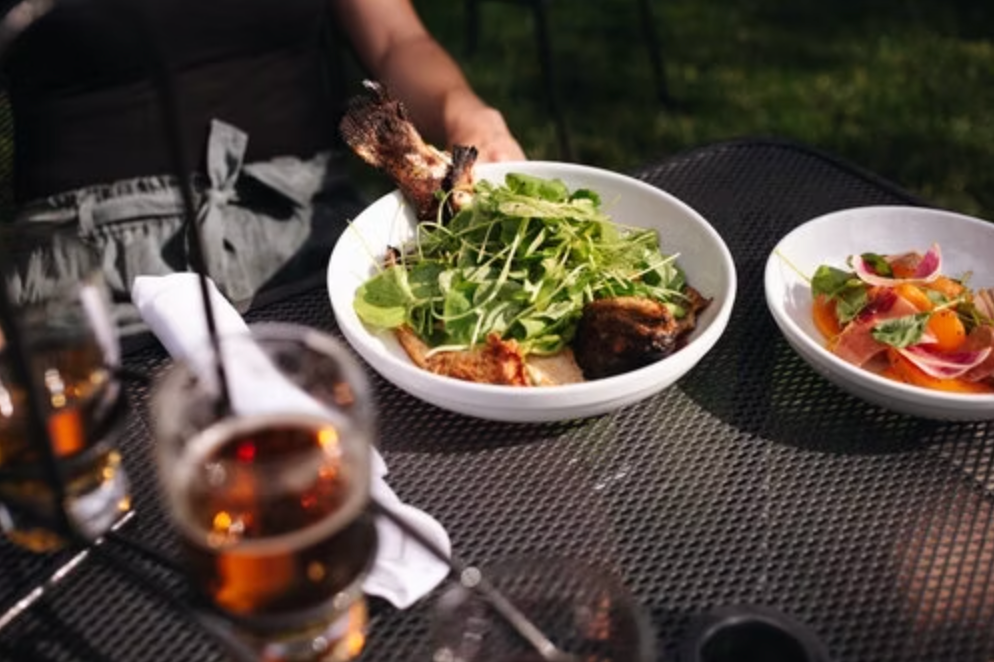 Outdoor dining table with white bowls of salad and greens, a glass of beer, and a person's hand holding a plate of food.