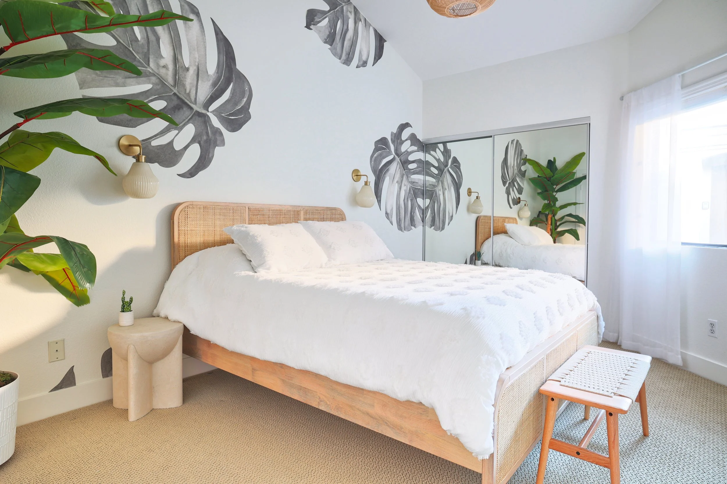 A cozy bedroom with a bed, white bedding, and a rattan headboard. The wall behind the bed features large black and white monstera leaf murals. There are two wall-mounted lamps above the bed, a small cow-shaped side table with a small cactus, and a mirrored closet door reflecting the room and some green plants. Sheer white curtains cover a window on the right side.