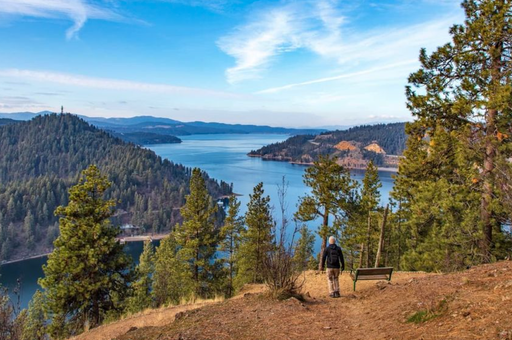 A hiker with a backpack walking along a trail overlooking a large blue lake surrounded by pine trees and rolling hills under a partly cloudy sky.