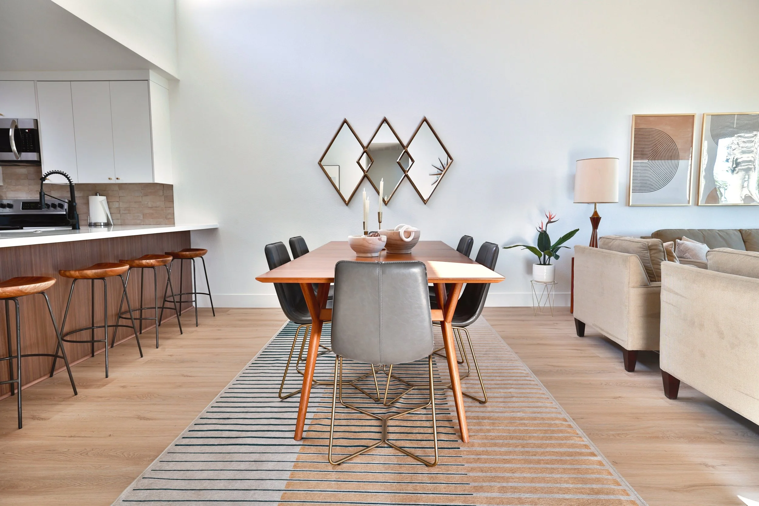 Modern dining area with a wooden table, six chairs, and a decorative mirror on the wall, adjacent to a patio with white kitchen and beige sofa.
