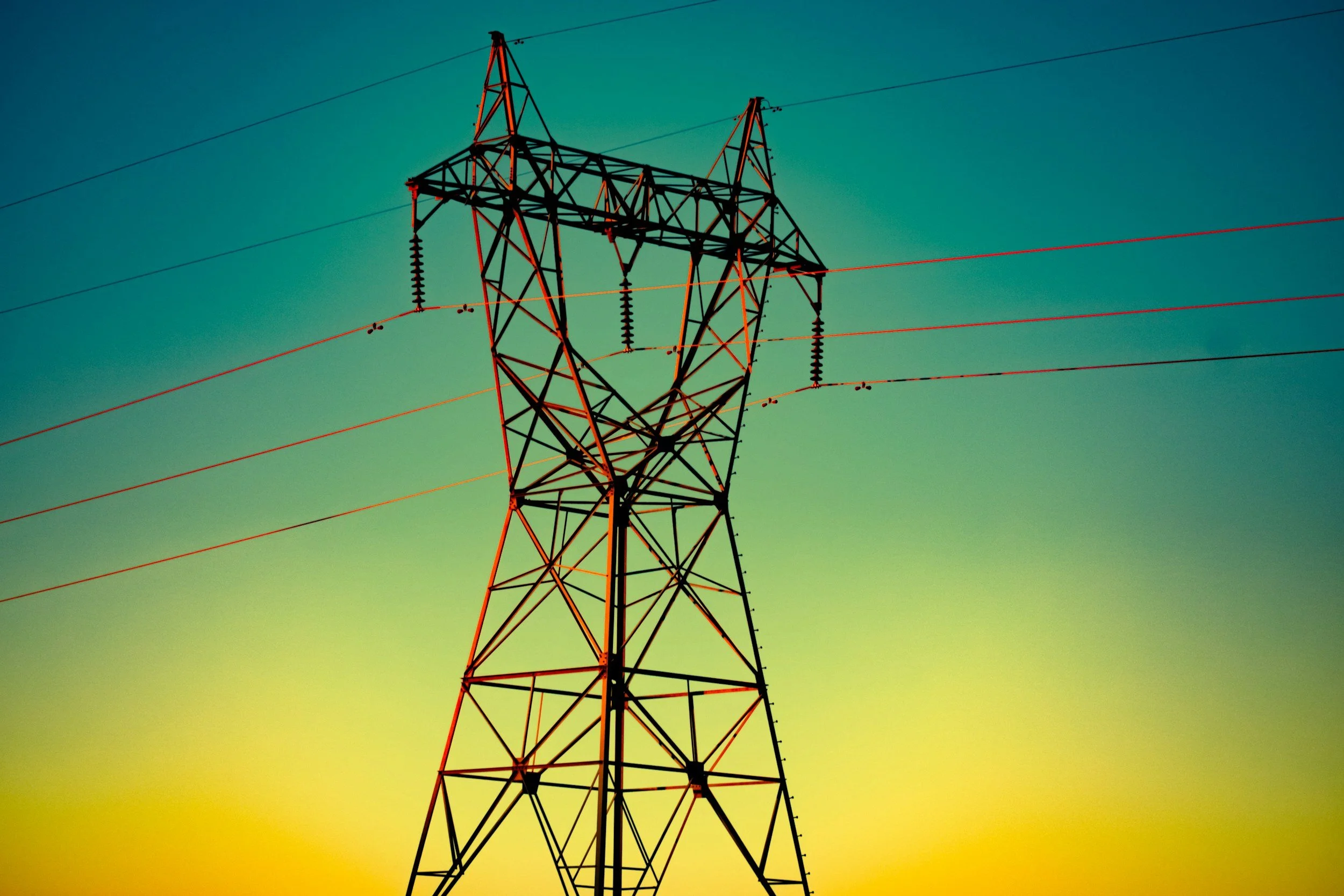 A power transmission tower silhouetted against a colorful sunset sky with green, yellow, and blue hues.