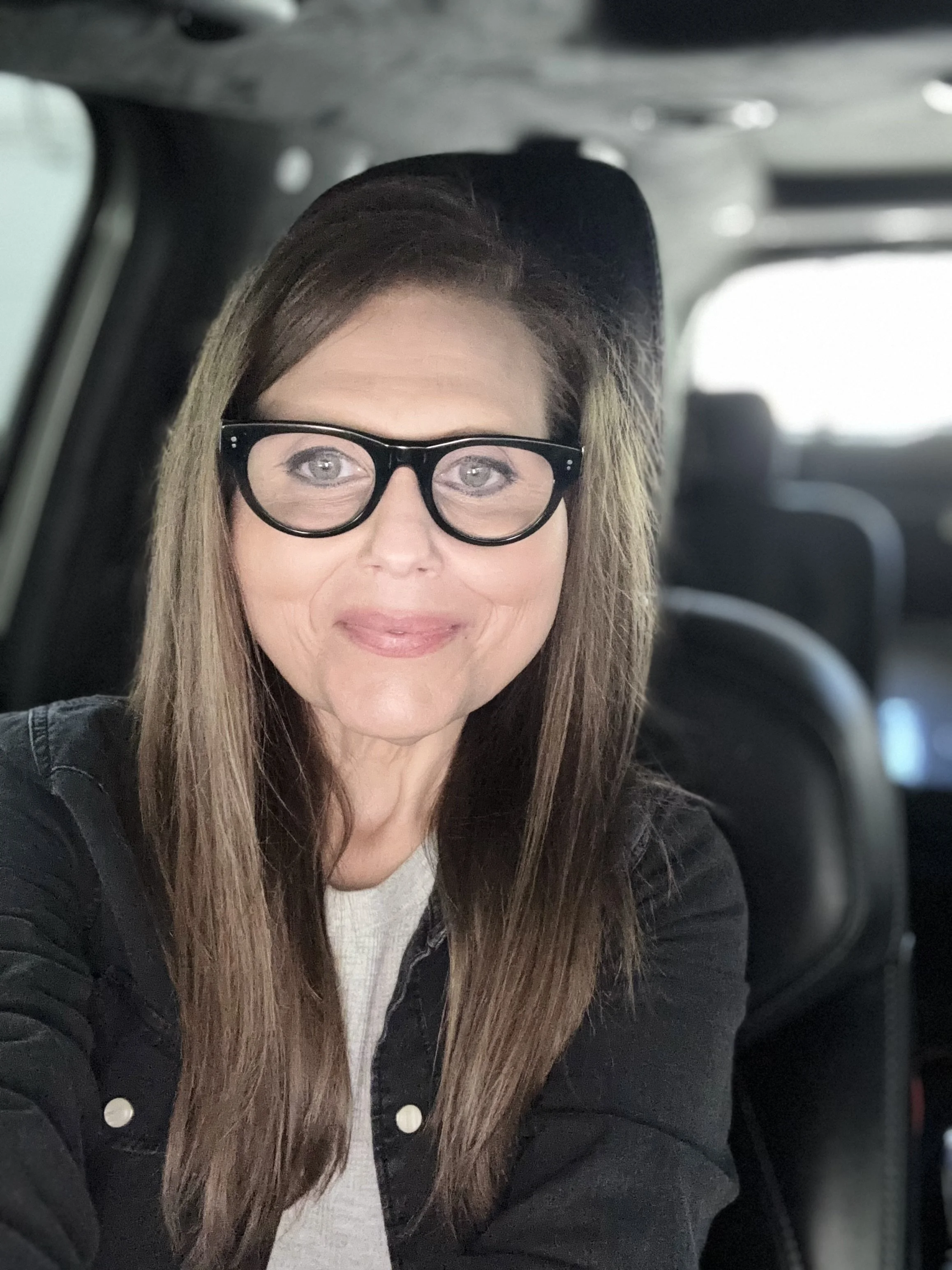A woman with long brown hair, glasses, and wearing a black jacket sitting inside a vehicle, smiling at the camera.