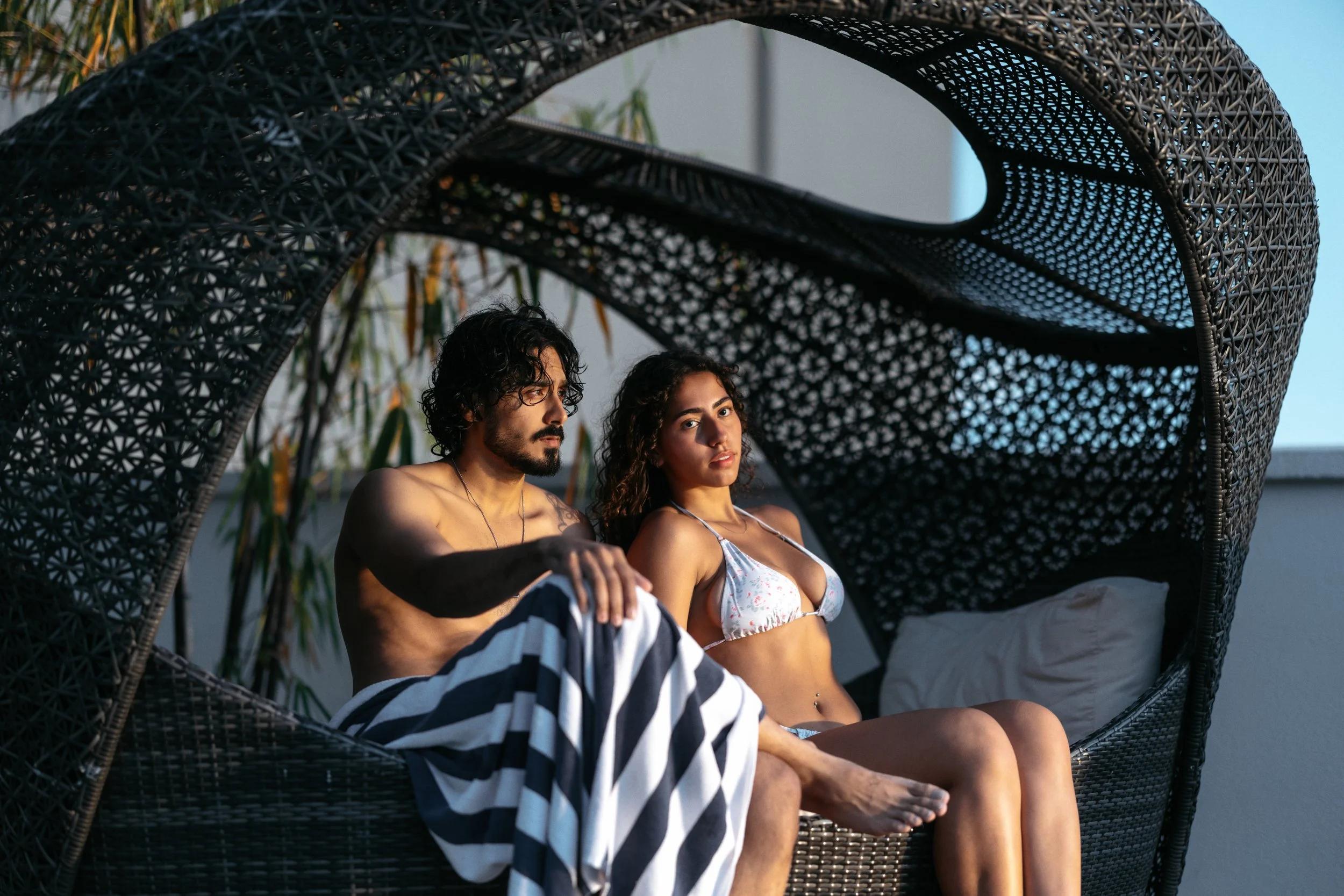 A man and woman sitting in a woven outdoor daybed with a canopy, at sunset.