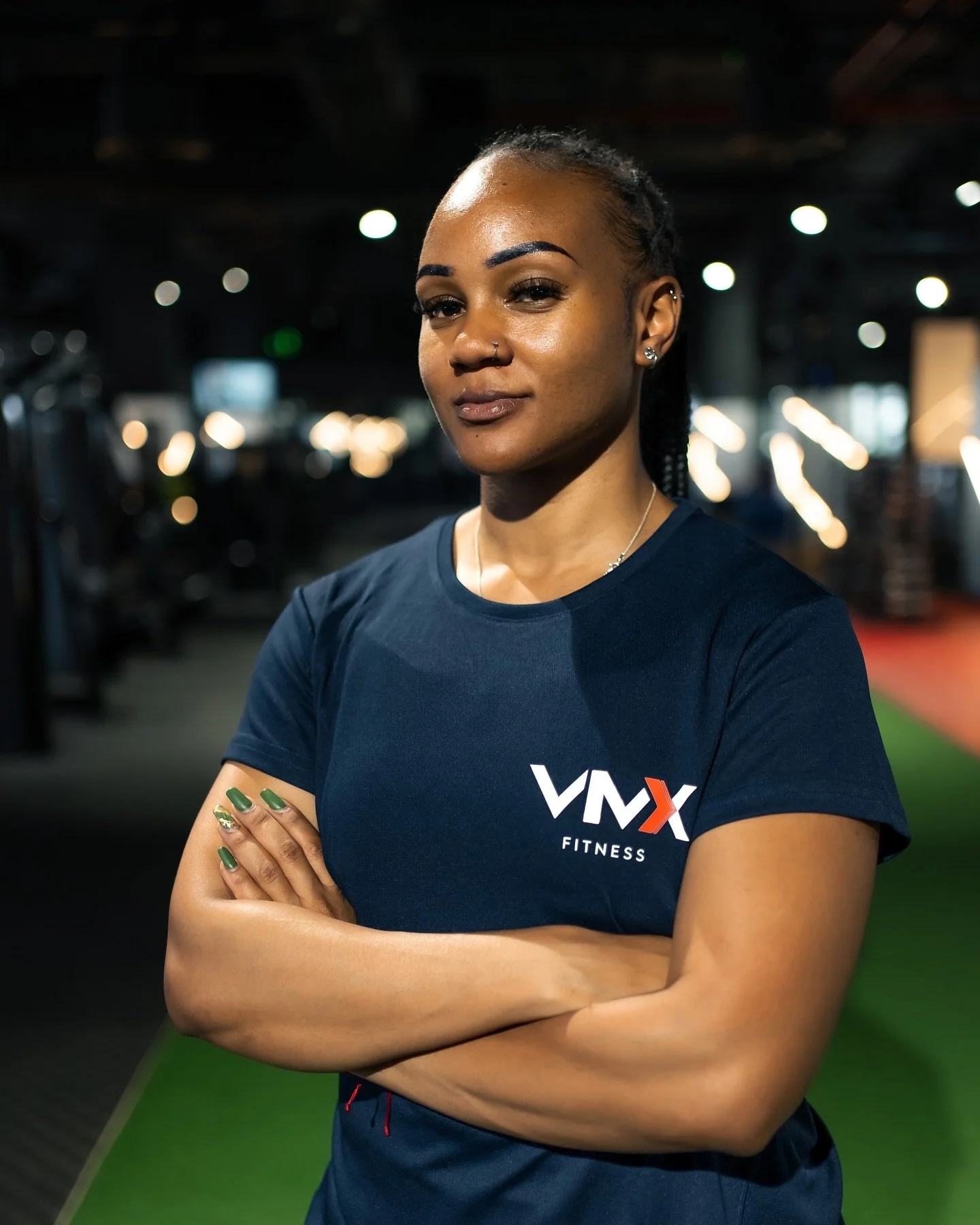 A confident woman in a dark blue fitness shirt with arms crossed, standing in a gym with blurred equipment and bokeh lights in the background.