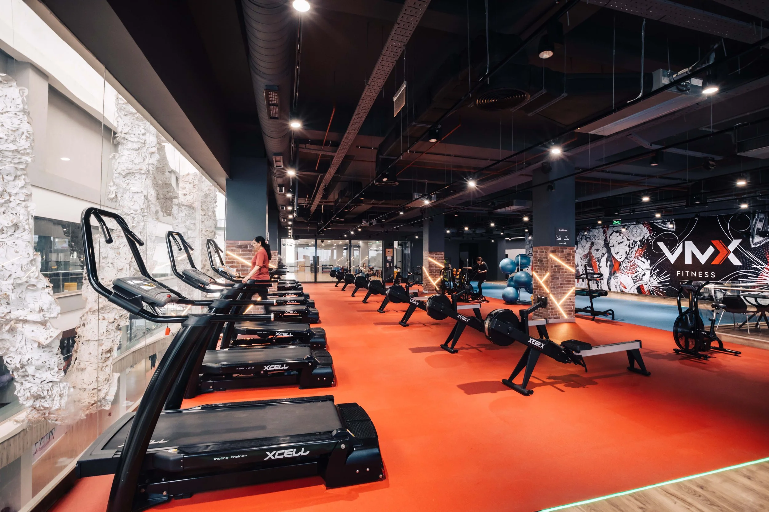 Modern fitness gym with cardio equipment including treadmills and rowing machines on a red floor. Large windows on the left, rock wall decor, and a wall with a fitness logo on the right. The gym is unoccupied except for one person using a treadmill.