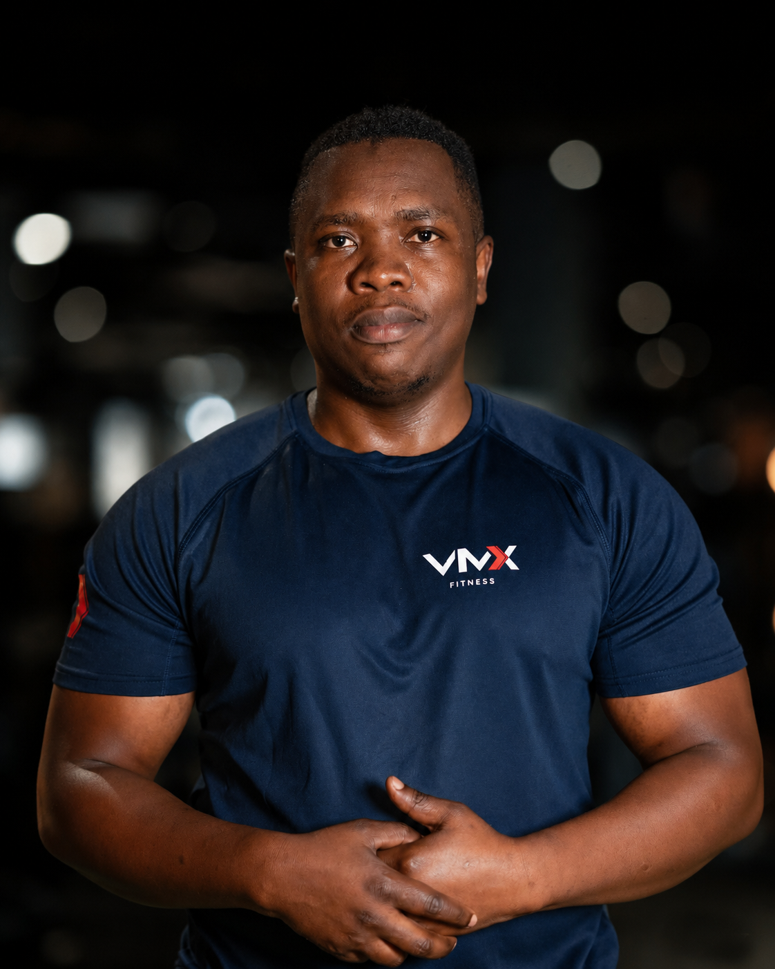 A person with short black hair in a navy blue shirt with a fitness logo, standing in a gym with workout equipment and other people in the background.