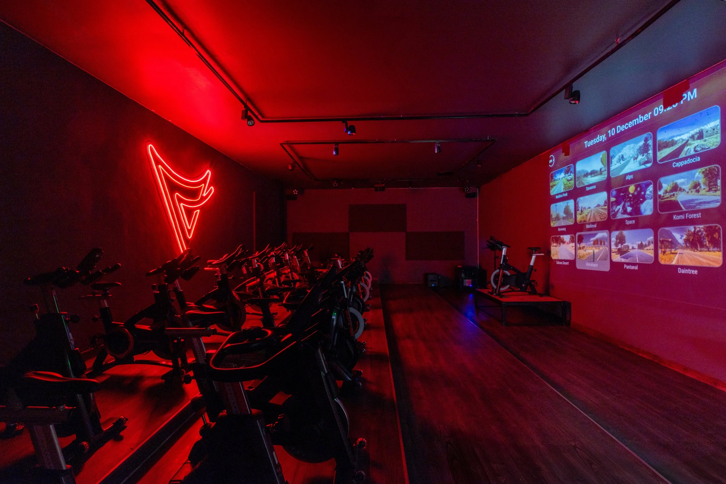 Indoor cycling studio with multiple exercise bikes illuminated by red neon light, with a large screen on the wall displaying various landscape images and the date, and a single stationary bike on a platform near the screen.